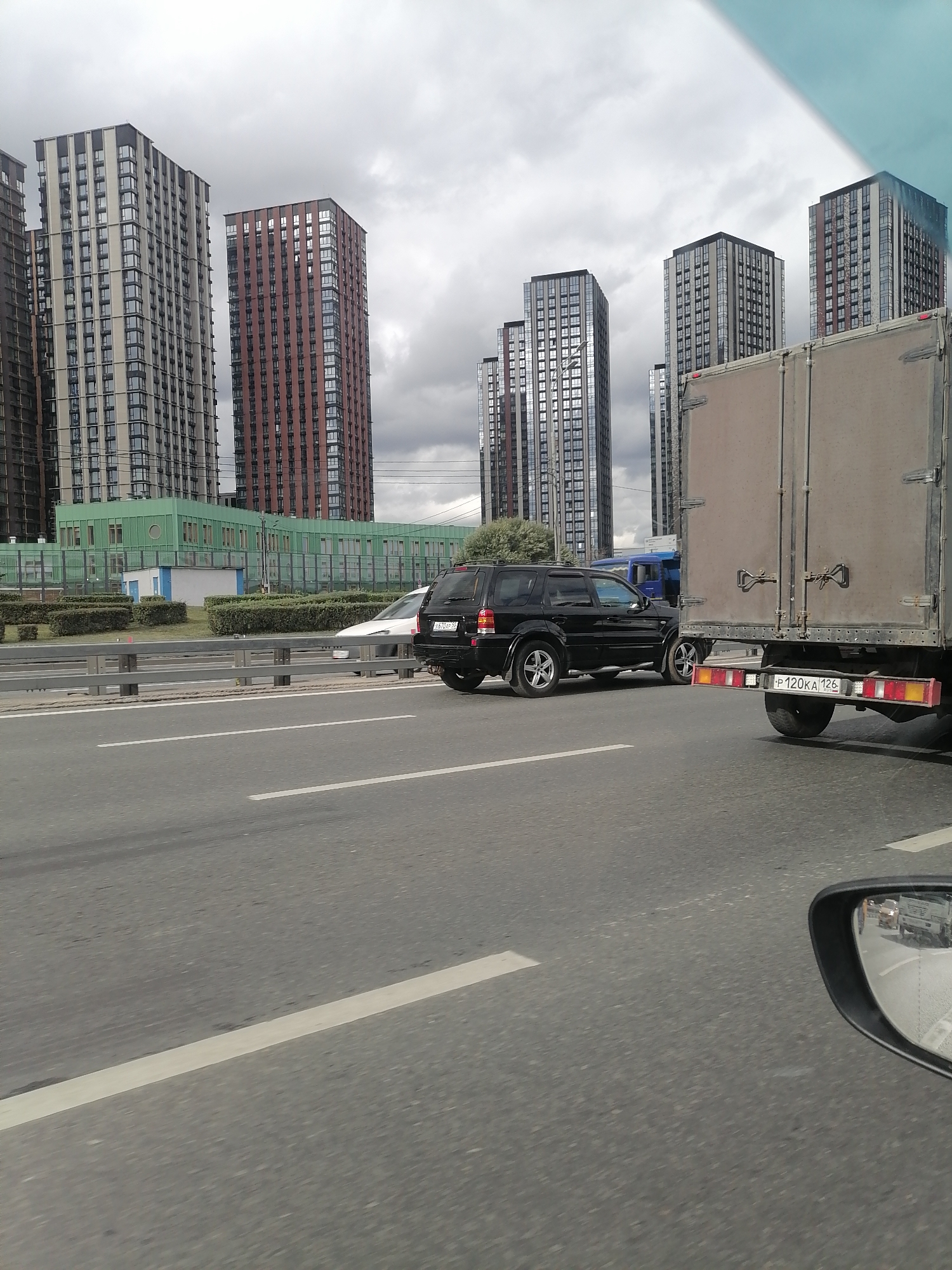 The image depicts a view from a moving vehicle on a highway, with a focus on a traffic scene and city skyline. In the foreground, the asphalt road surface is visible with white lane markings. A black SUV is driving in the middle lane, and a large cargo truck is in the right lane. The truck's rear license plate is visible, reading "P 120KA 126".
In the background, a row of modern high-rise residential buildings with a mix of beige, red, and dark-colored facades dominates the cityscape. A green administrative building with a circular feature is situated in front of some of the residential towers. A light-colored sedan is visible in the left lane. The sky is overcast with dense clouds, indicating a cloudy day. A side mirror of the vehicle from which the photo is taken reflects other vehicles and a portion of the road.
The scene is characteristic of a suburban or outer district of a large city with significant new residential development. The architecture of the buildings, the presence of a highway, and the license plate format suggest a location in Russia, possibly on the outskirts of Moscow, Russia. The overall atmosphere is one of urban infrastructure and daily commute under a muted sky.
