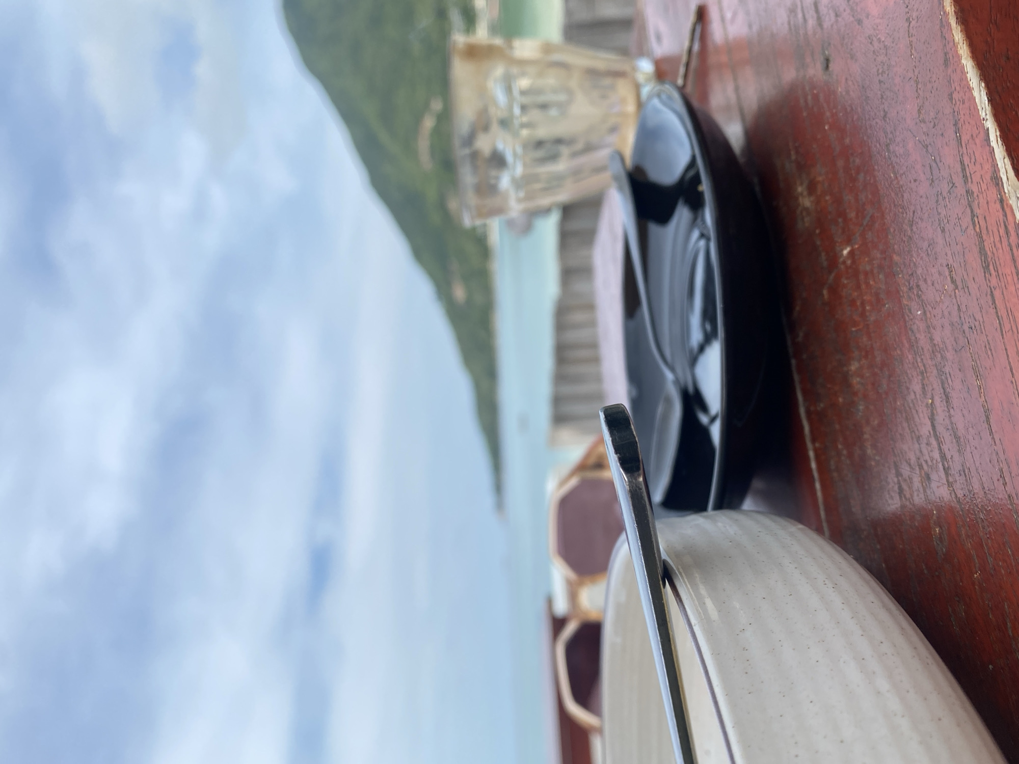 On a weathered, dark red wooden table sits a white speckled bowl with a silver spoon resting inside, a black bowl partially obscuring a clear glass, and a pair of sunglasses. In the background, a view of Baan Chaloklum, Thailand can be seen, featuring a body of water, green hills, and a cloudy sky.