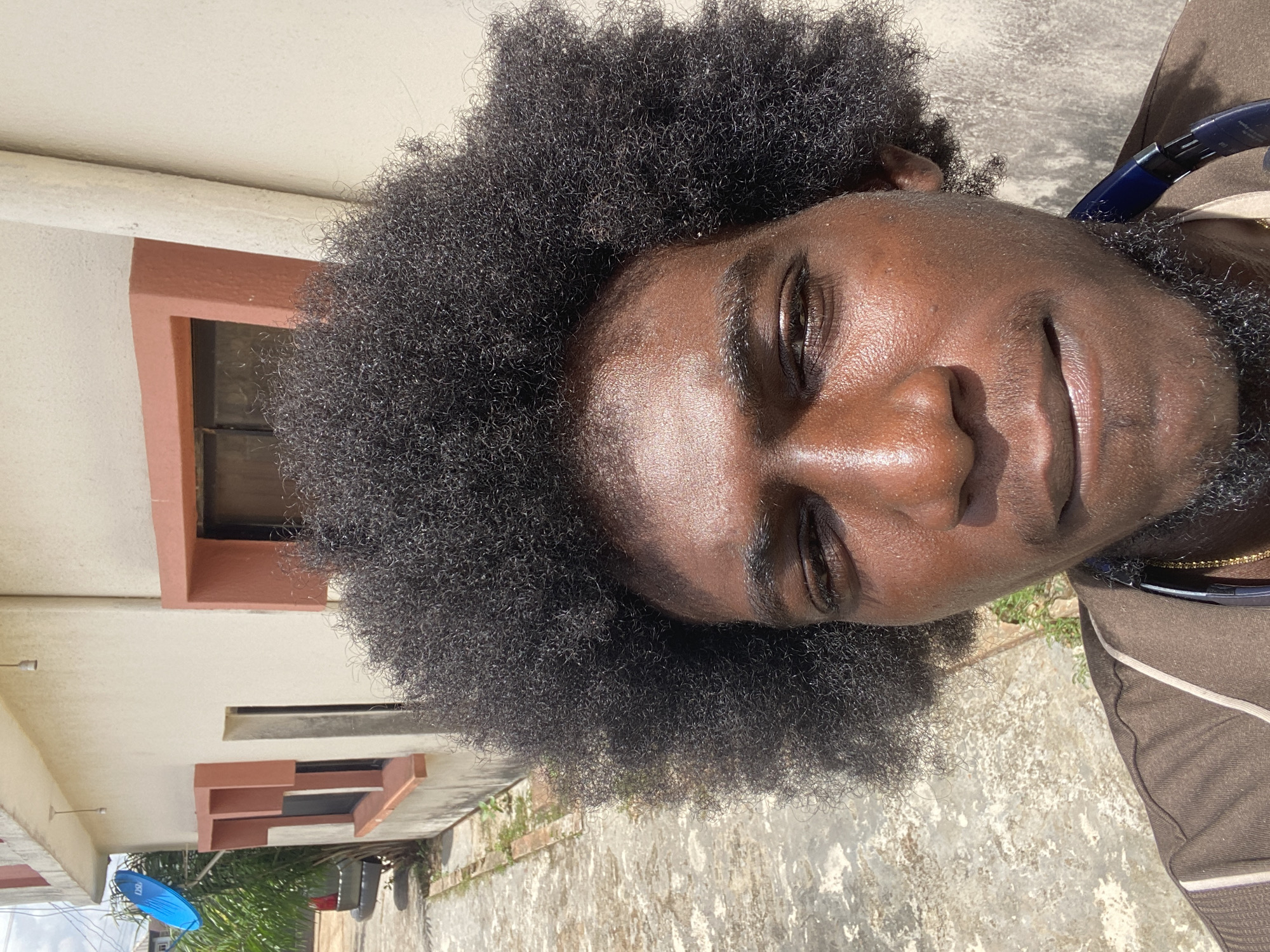 A close-up, slightly rotated shot captures a Black man with a short beard and dark, Afro-textured hair. He is facing the camera and has a slight smile. He is wearing a brown shirt with white trim. There is a blue object in the bottom left, and a building with windows.
While specific identification of the location in Lagos, Nigeria, is impossible without additional information, the presence of a building and an outdoor setting suggests an urban or suburban environment.