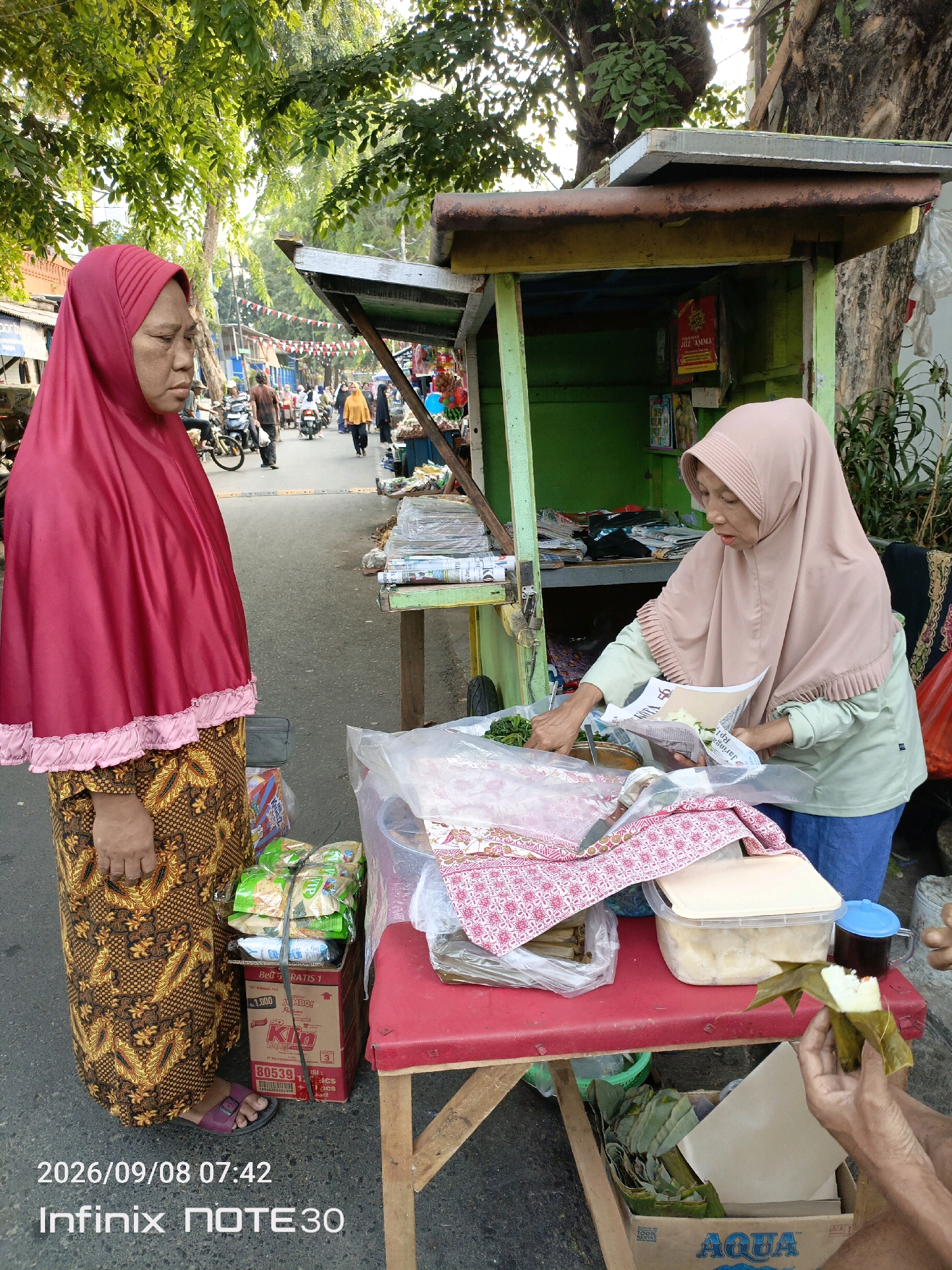The image captures a street scene in Central Jakarta, Indonesia, on September 8, 2026, at 7:42 AM. Two women are the primary subjects. The woman on the left, wearing a maroon hijab and a patterned skirt, stands facing away from the viewer, looking towards the street. The woman on the right, wearing a beige hijab and a light green shirt, is engaged with merchandise at a roadside stall. She is holding a piece of paper, possibly a receipt or product information, and appears to be interacting with the stall's offerings.
The stall itself is simple, with a wooden frame and a green-painted structure. It displays various items, including packaged goods, what appears to be food items in plastic containers, and some leafy green vegetables. A red-covered table in front of the stall holds more merchandise. In the foreground, a person's hands are visible, unwrapping a food item wrapped in banana leaves.
The background shows a street with other people, motorcycles, and more stalls, suggesting a bustling market or commercial area. Red and white triangular flags are strung overhead, adding a festive or decorative touch to the scene. The overall environment indicates a typical urban setting in Indonesia, likely a busy street market during the morning hours.