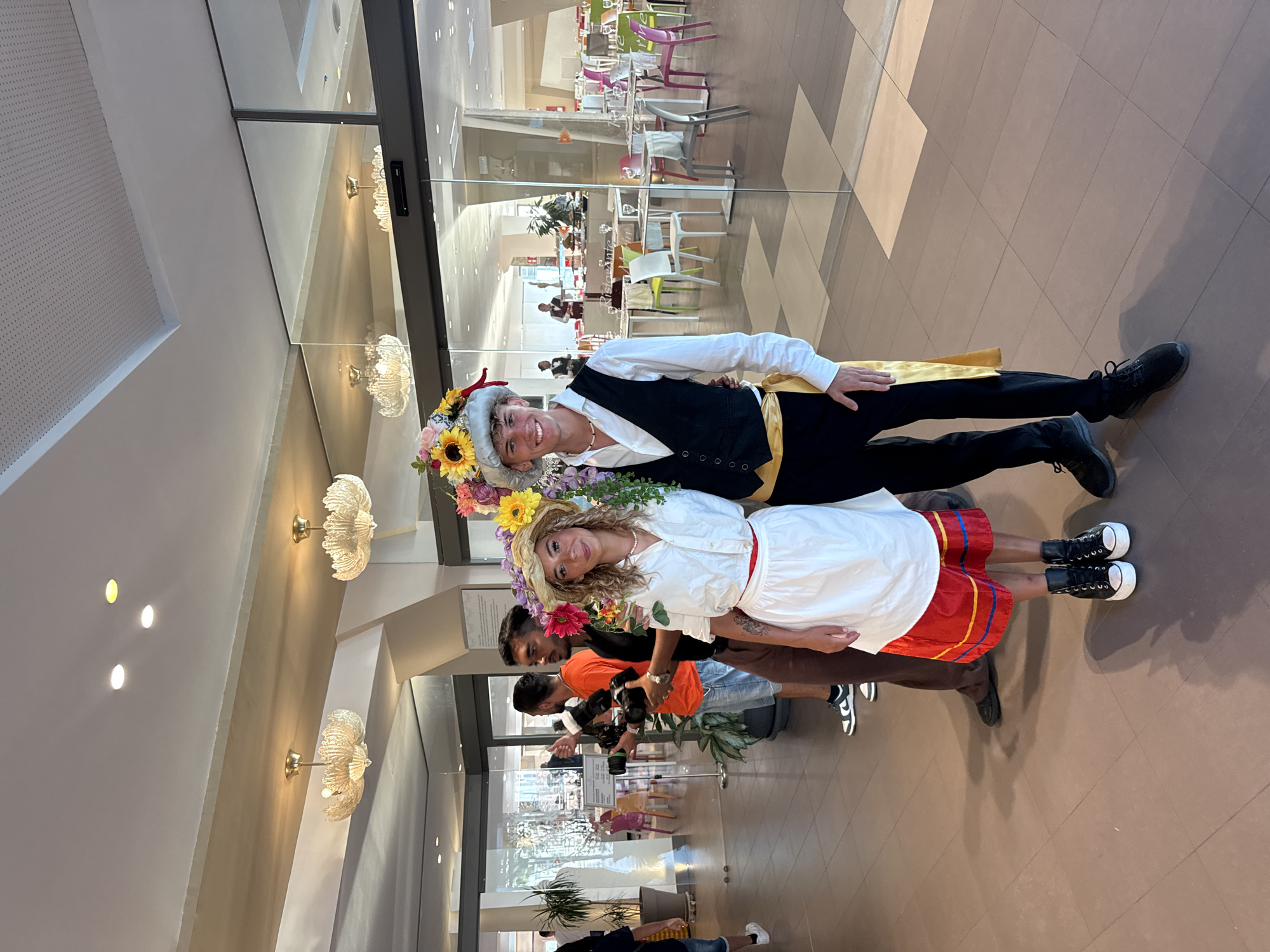 The image shows a young couple dressed in traditional attire standing in an indoor space. The woman wears a white shirt, a white skirt with a red waistband and a red and yellow trim, and black high-top sneakers. She also has a straw hat adorned with flowers. The man wears a white shirt, a black vest, black pants, a yellow sash, and black sneakers. He also has a decorative hat with flowers and other embellishments. They are standing in front of a glass-partitioned area, which appears to be a restaurant or dining hall with tables and chairs visible in the background. The lighting suggests it is daytime, possibly late afternoon. The floor is tiled with a pattern of brown and beige squares. In the background, other people are visible, some appearing to be taking photos. This scene is not specifically identifiable as being in the city of Isola delle Femmine, Italy.