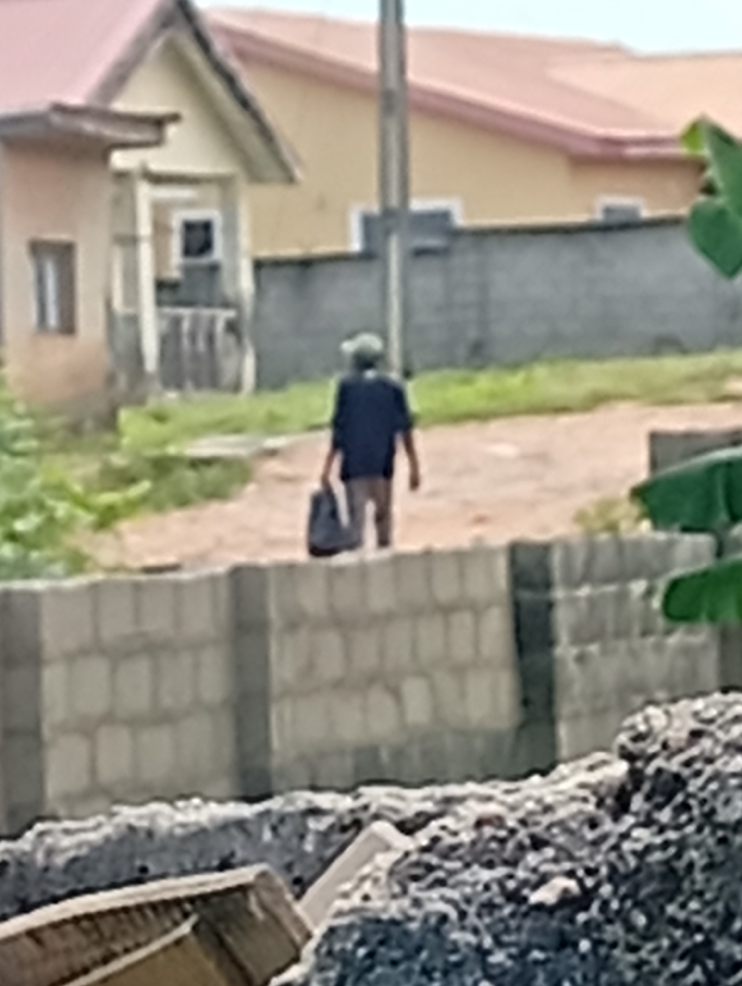 A person wearing a dark shirt and shorts walks away from the camera in an outdoor environment. The person is carrying a dark bag in their right hand. The person is walking on a dirt path. In the background, there are buildings with red and yellow roofs and a gray brick wall. There is also some green vegetation. In the foreground, there is a low brick wall and a pile of debris. The scene appears to be in an urban or suburban area.