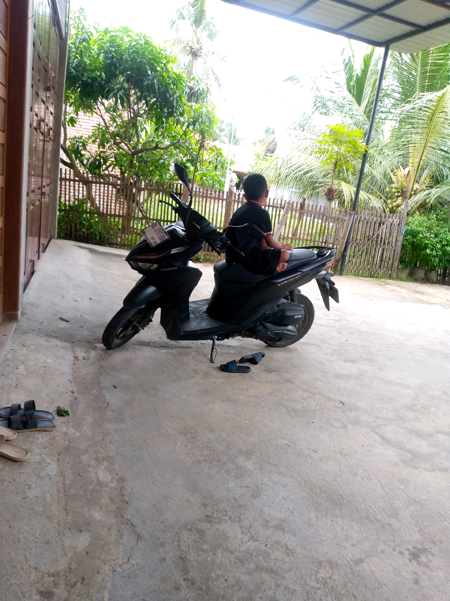A child sits on a black Honda scooter in an outdoor area with a concrete floor, possibly a yard or driveway. The child, wearing a black t-shirt and shorts, faces away from the camera, looking towards a backdrop of lush green trees and palm trees. A bamboo fence is visible behind the scooter and trees. The area appears to be in Ujong, Indonesia, suggested by the tropical vegetation. In the foreground, a pair of black flip-flops and another pair of lighter-colored sandals lie on the concrete. A portion of a building with brown wooden doors is visible on the left side of the image.