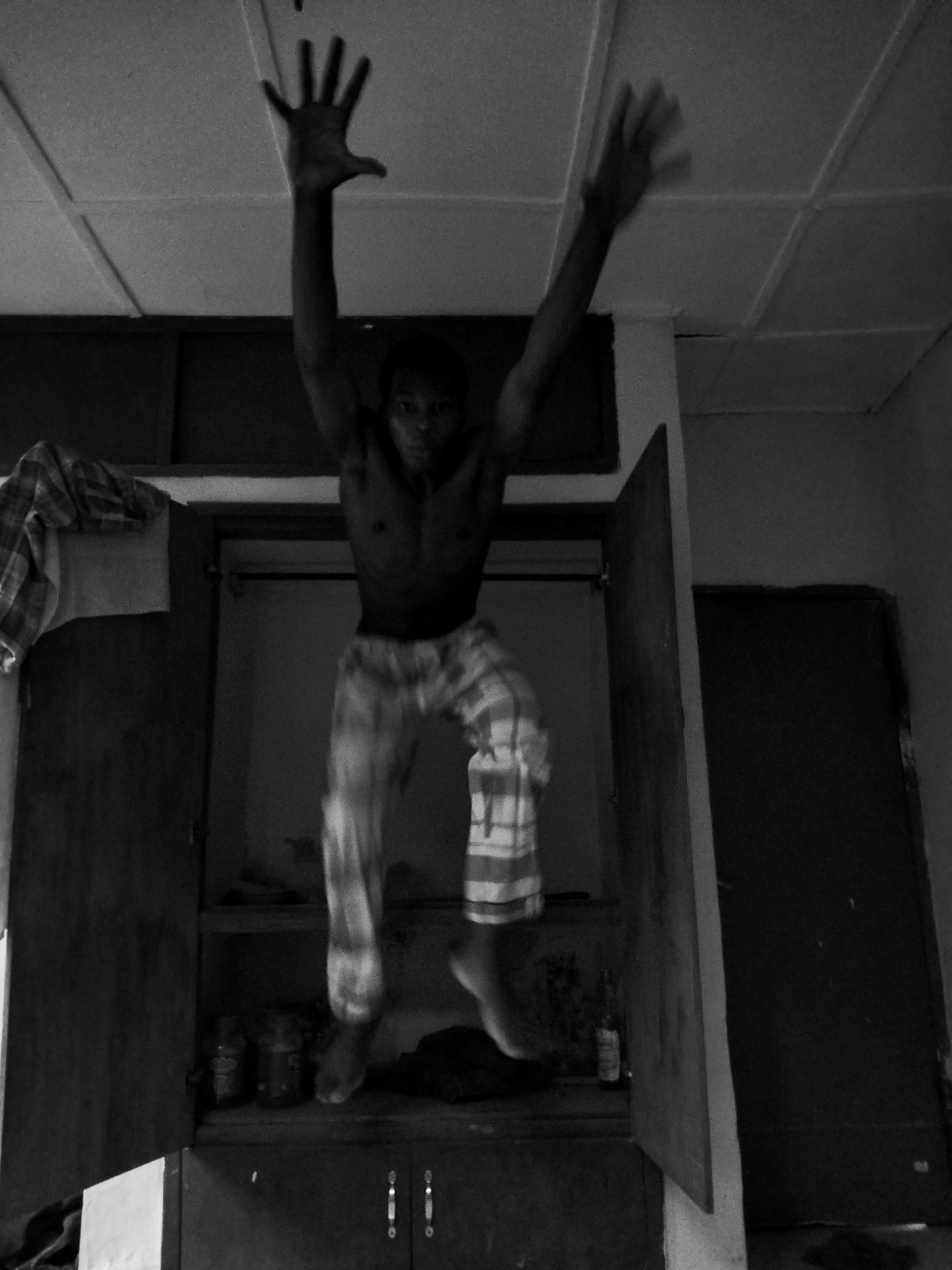 A black-and-white photo depicts a person jumping inside an open closet. The person, shirtless and wearing patterned pants, has both arms raised. The setting appears to be indoors, likely a room with a tiled ceiling. The closet contains shelves with various jars and bottles. A door is visible to the right of the closet. The location is not specified in the image itself. Therefore, the location in Kubwa, Nigeria, cannot be confirmed from the image alone.