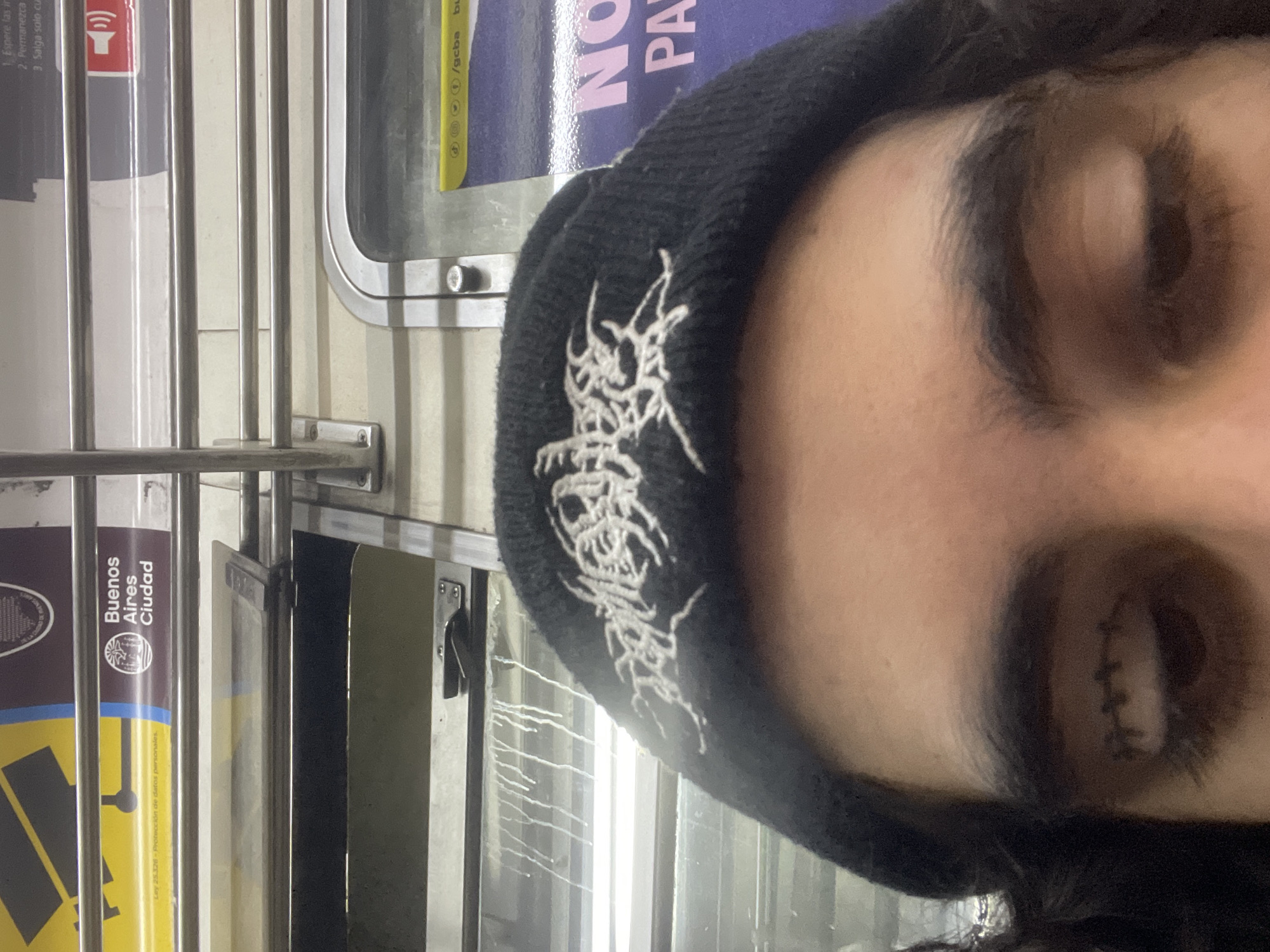 A close-up, slightly angled shot features a person wearing a black beanie with white gothic-style lettering, partially obscuring their forehead and the top of their face. The person's eyes are closed. The background reveals elements of public transportation, identifiable as a train or subway car in Buenos Aires, Argentina. This is indicated by the signage visible on the left side of the frame, which includes the phrases "Buenos Aires Ciudad" and mentions the "Ley 25.326 - Protección de datos personales." Further signage shows the website address "/gcba" and partial words such as "NO" and "PA." Vertical metal bars and the edge of a window are visible, adding to the context of being inside a public transit vehicle.