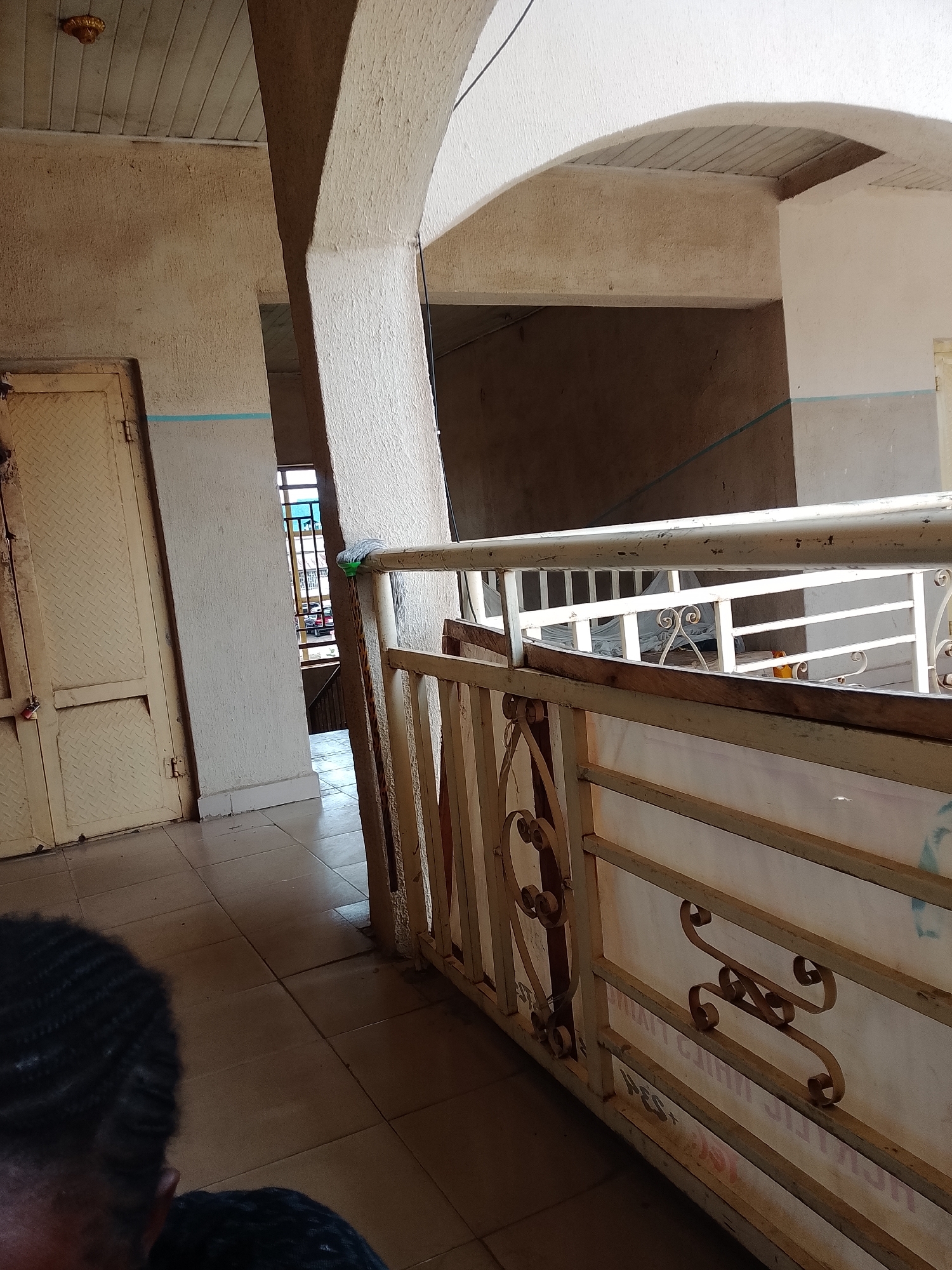 This is a medium shot, eye-level, outdoor photograph taken in Jalingo, Nigeria. The image appears to be taken during the daytime, with no indication of specific weather conditions.
The foreground shows the back of a person's head and shoulders, with dark, curly hair. The person is looking away from the camera, towards a balcony or walkway.
The midground features a railing with ornate metalwork and wooden accents. Beyond the railing, an arched passageway leads to a staircase. The walls are textured and painted in neutral tones. A yellow door with a diamond pattern is visible on the left.
The background shows a glimpse of a street or courtyard with cars, indicating an urban or semi-urban setting. There is some visible text on a banner or sign behind the railing, but it is largely obscured and illegible. The overall impression is one of a utilitarian building or public space, possibly an apartment complex or a commercial establishment. The lighting suggests it is daytime, and the ambient light is somewhat muted.