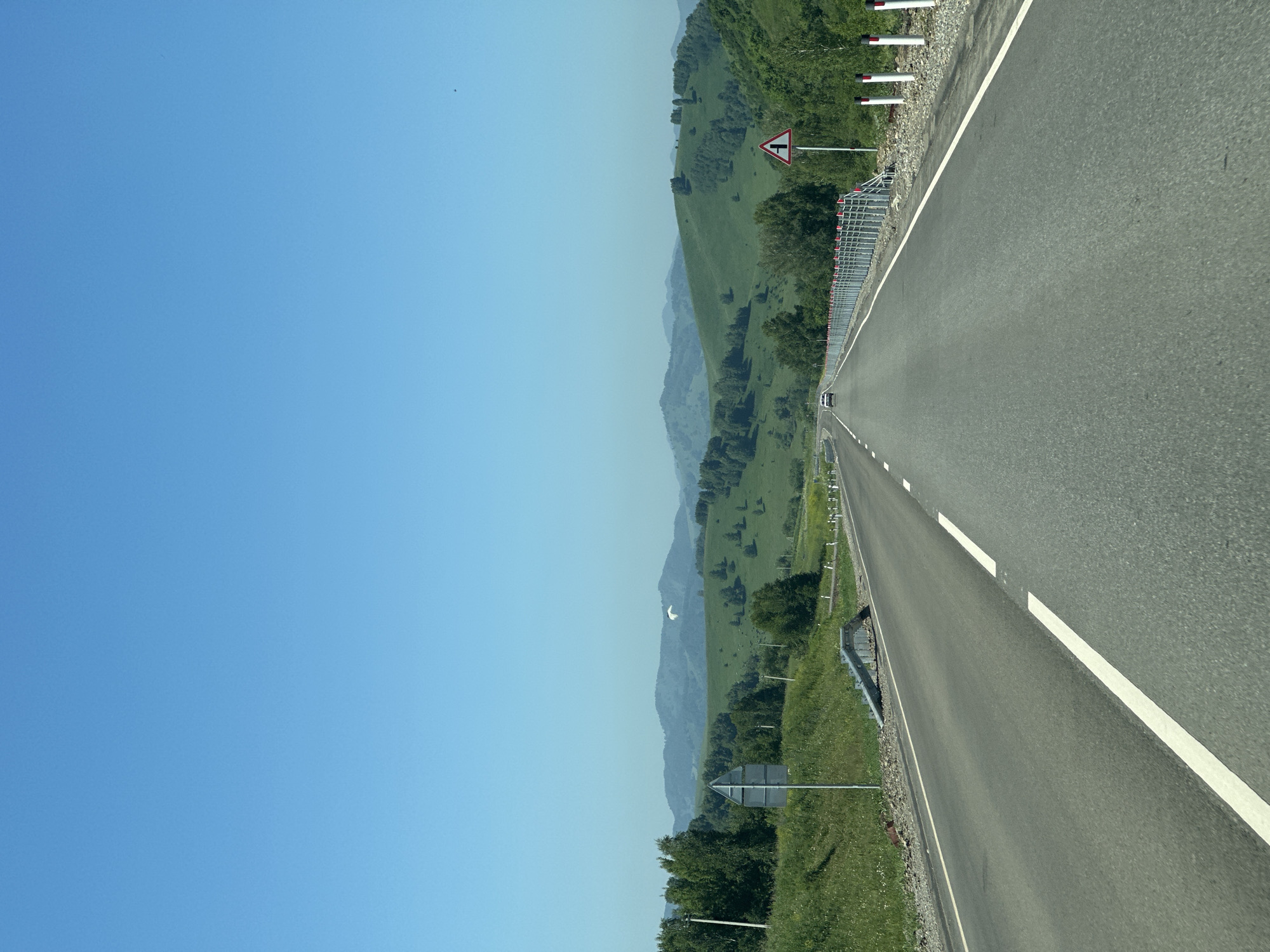 This image depicts a clear, sunny day on a paved road in Verkh-Aya, Russia. The road stretches into the distance, bordered by green, rolling hills covered in trees. A white double solid line marks the center of the road, with a white dashed line to the right. A guardrail with red and white reflective posts runs along the right side of the road. Several traffic signs are visible, including a triangular warning sign indicating a junction on the right. A white car is visible in the distance, traveling away from the camera. A single white bird is flying in the sky. The sky is a bright, clear blue.