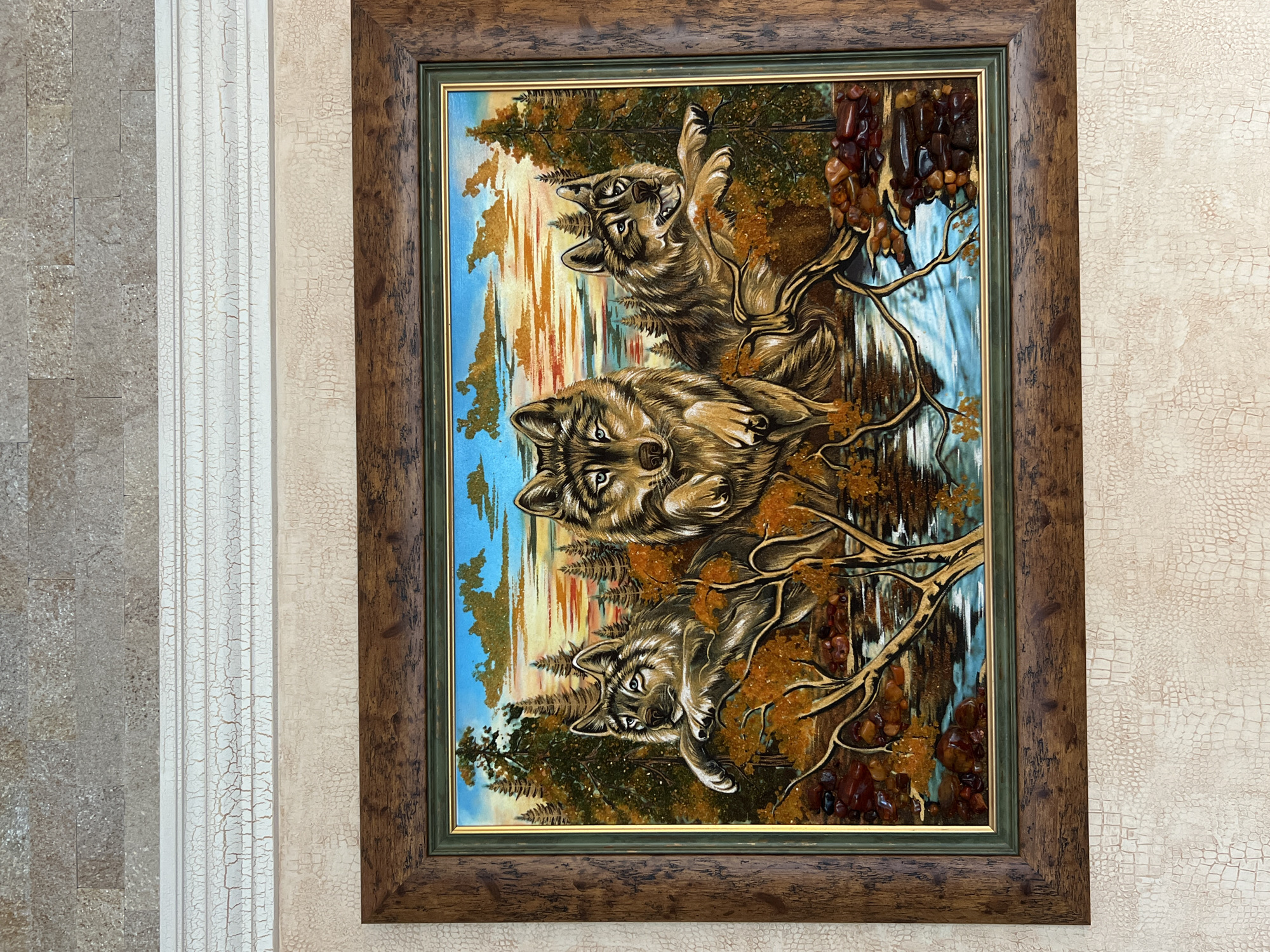A framed painting of three wolves is displayed on a textured wall. The painting depicts the wolves in a forest setting with a river and a sunset in the background. The wolves are rendered in shades of brown and gray, with piercing blue eyes. The leaves on the trees appear to be made of amber. The painting is framed in a dark wooden frame with a green mat. The wall behind the painting is covered with a textured wallpaper. The location is not specified in the image.