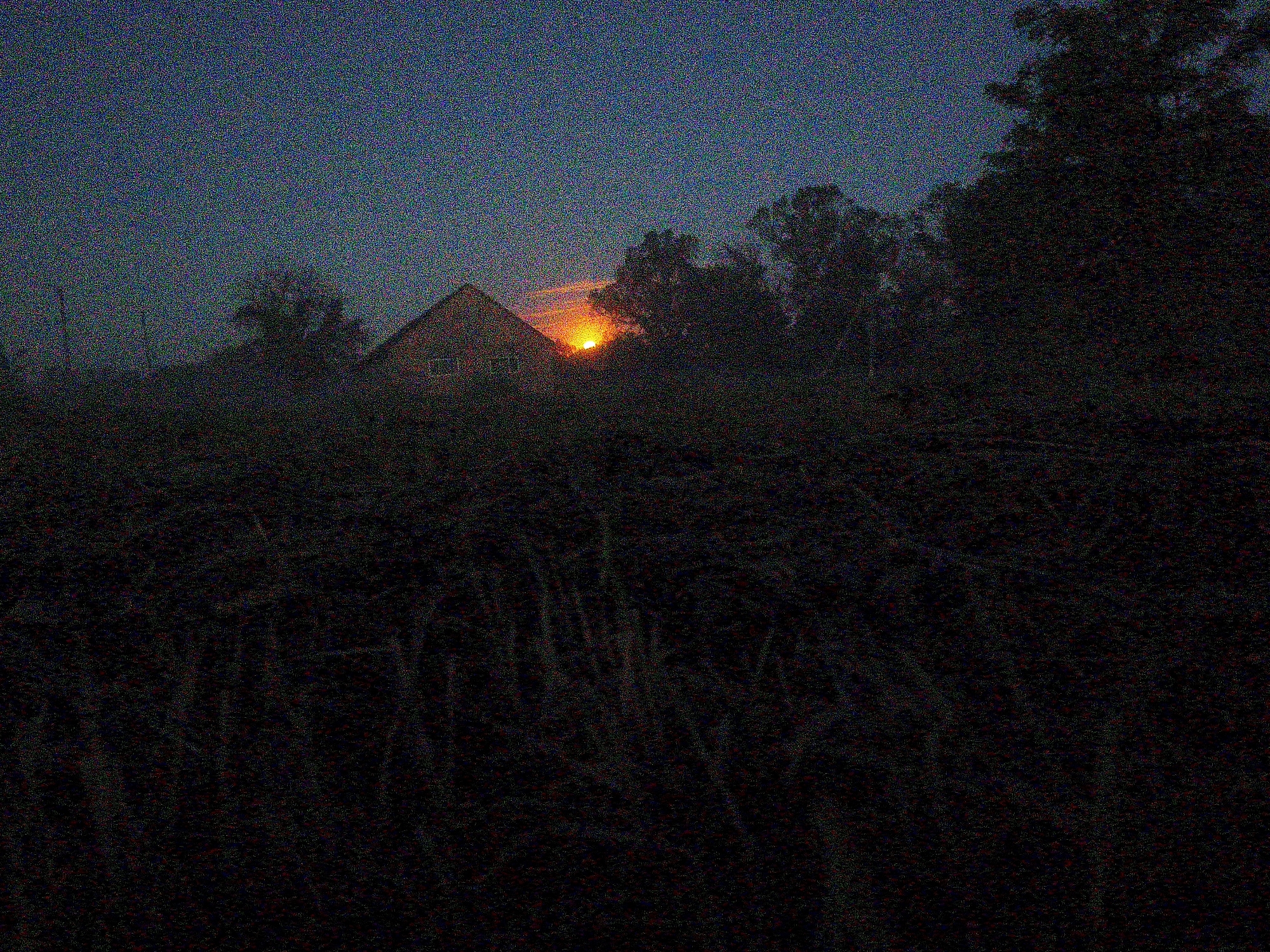 The image depicts a rural scene at dusk or dawn. A building, possibly a house, is visible in the middle ground, silhouetted against a bright orange glow on the horizon, likely from the rising or setting sun. Trees and vegetation surround the building. The foreground is dark, filled with dense foliage, obscuring details. The sky is a dark blue or grey, suggesting twilight. The high levels of noise and the lack of details makes it hard to pinpoint the exact location, people and activities. I do not have enough information to accurately identify specific location in Ustyvytsia, Ukraine.