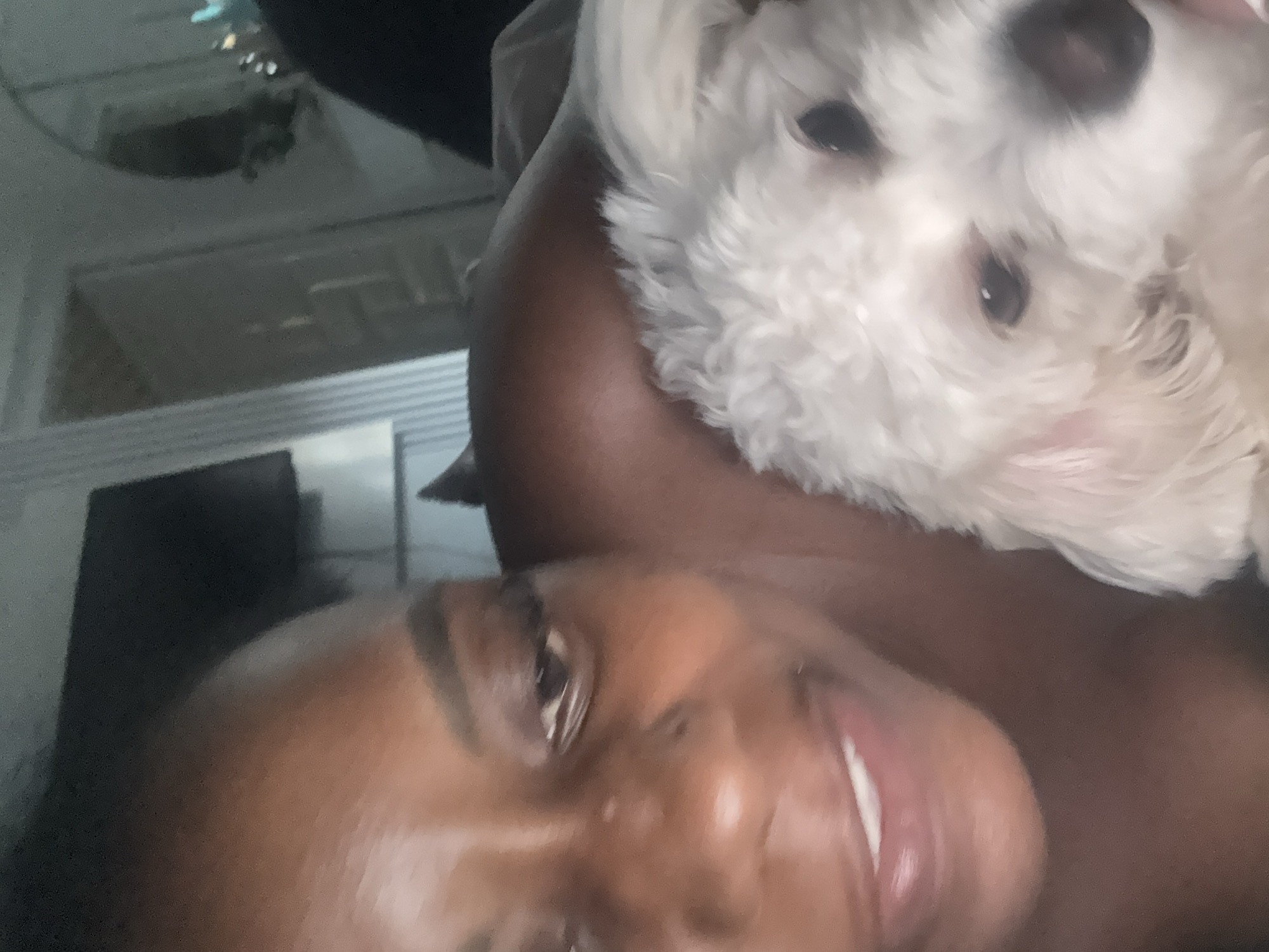 The image shows a close-up of a woman and a white dog. The woman, who appears to be of African descent, is looking at the camera with a slight smile. She has dark hair and is wearing makeup. The dog is a small, fluffy white breed, possibly a Bichon Frise or Poodle mix, with dark eyes and a dark nose. The dog is resting its head on the woman's shoulder or chest. The background is blurred but suggests an indoor setting with some furniture and a wall with vertical lines. The lighting is dim, indicating it might be evening or a dimly lit room. The overall impression is an intimate moment between a woman and her pet. There is no information or visual cues in the image to suggest a specific location in Abuja, Nigeria.
