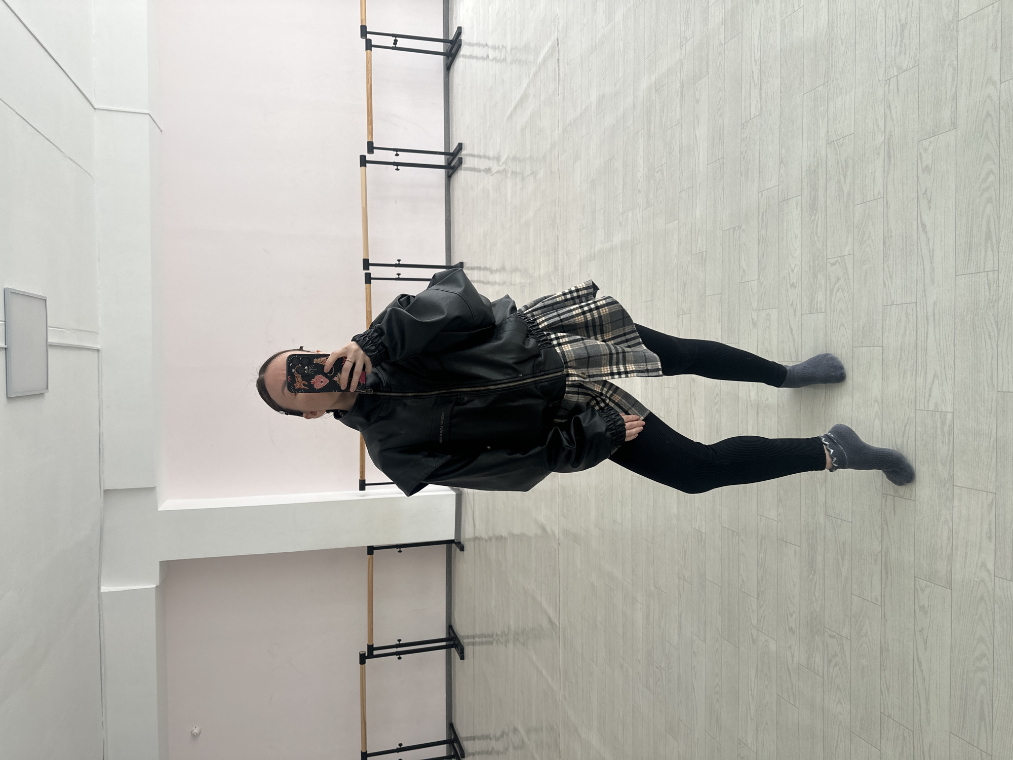 The image shows a person indoors, likely in a dance or exercise studio in Votkinsk, Russia. The person is standing on a light-colored, wood-patterned floor and appears to be taking a selfie with a phone. They are wearing a black leather jacket, a plaid skirt, black leggings, and gray socks. In the background are ballet barres attached to the wall, and the walls are a combination of white and light pink. A rectangular object is visible on the left wall.