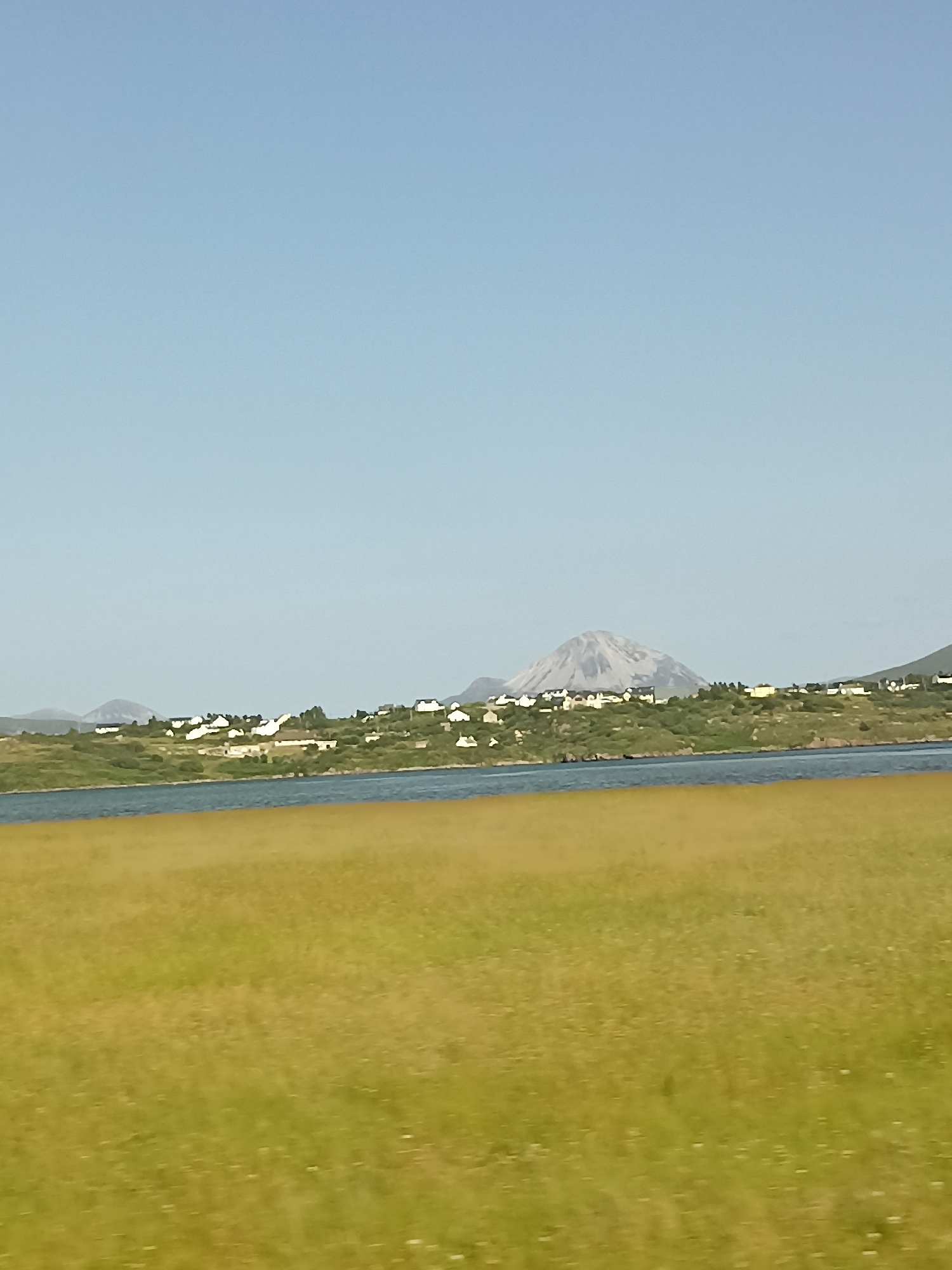 A wide-angle, landscape photograph shows a tranquil scene with a clear blue sky dominating the upper portion. Below the sky, a distant, conical mountain with a pale, rocky surface is visible, its peak partially shrouded in a light mist or cloud. In front of the mountain, a lush green hillside slopes down towards a body of water. Dotted along the hillside are numerous white-roofed houses, suggesting a small village or settlement. The water, a deep blue, separates the hillside from a foreground of tall, dry, golden-brown grass, which appears to be a meadow or field. The overall impression is one of a remote, peaceful, and scenic coastal or lakeside location, possibly in a rural area of Ireland like Ranafast, known for its natural beauty and scattered settlements.