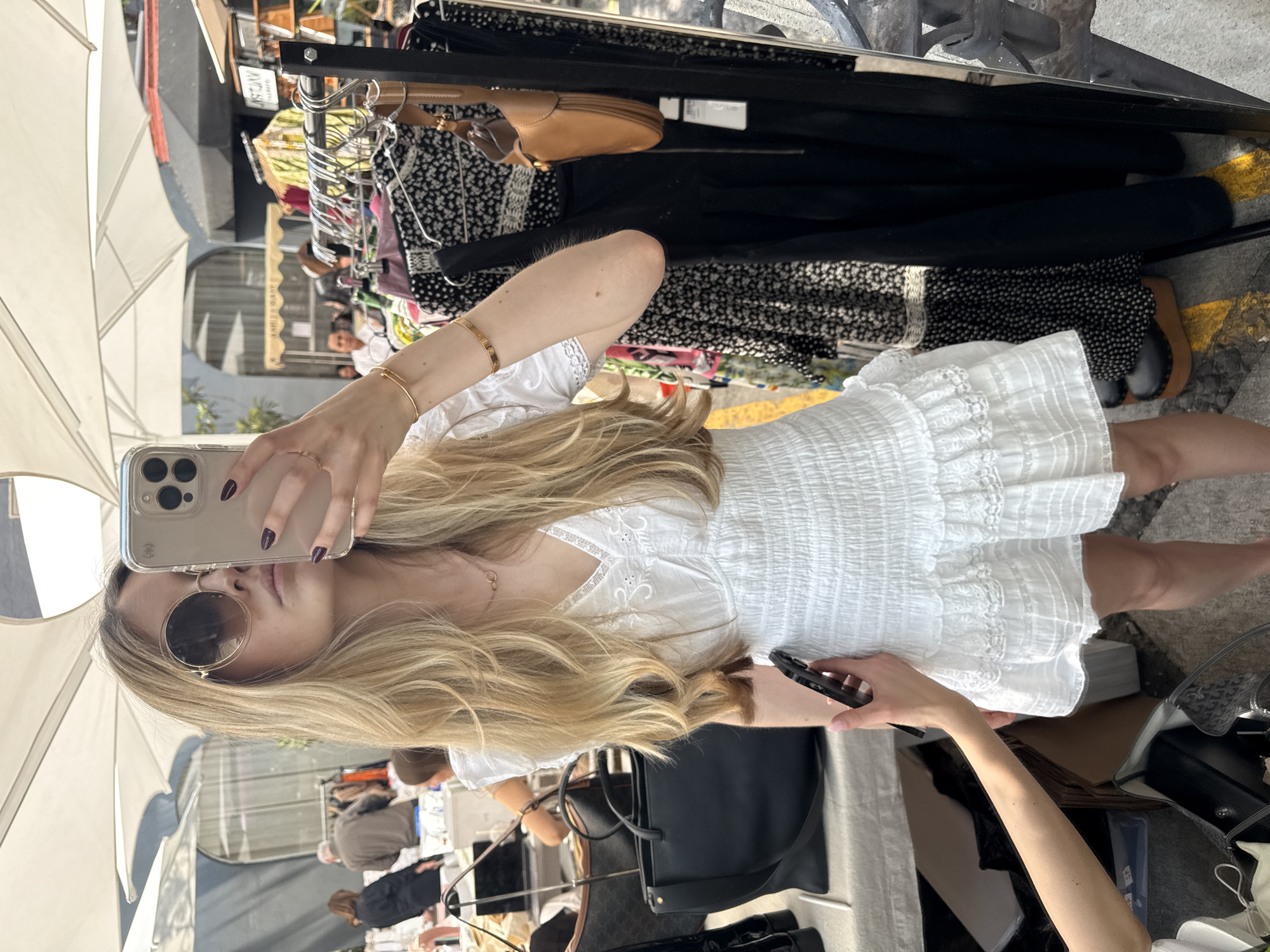 A woman is taking a mirror selfie in what appears to be a market or outdoor shopping area. She is holding her phone in her left hand and looks towards the camera. She is wearing sunglasses, a white dress with a ruffled skirt, and several gold bracelets on her left wrist. Her long, blonde hair falls over her shoulder and down her chest.
The background contains clothing racks filled with garments, handbags, and other items for sale. People are visible in the background, suggesting a busy market scene. The presence of an awning overhead indicates that the setting is outdoors.
Based on the visible elements in the photo, it is difficult to confirm that this picture was taken in Mexico City, Mexico.