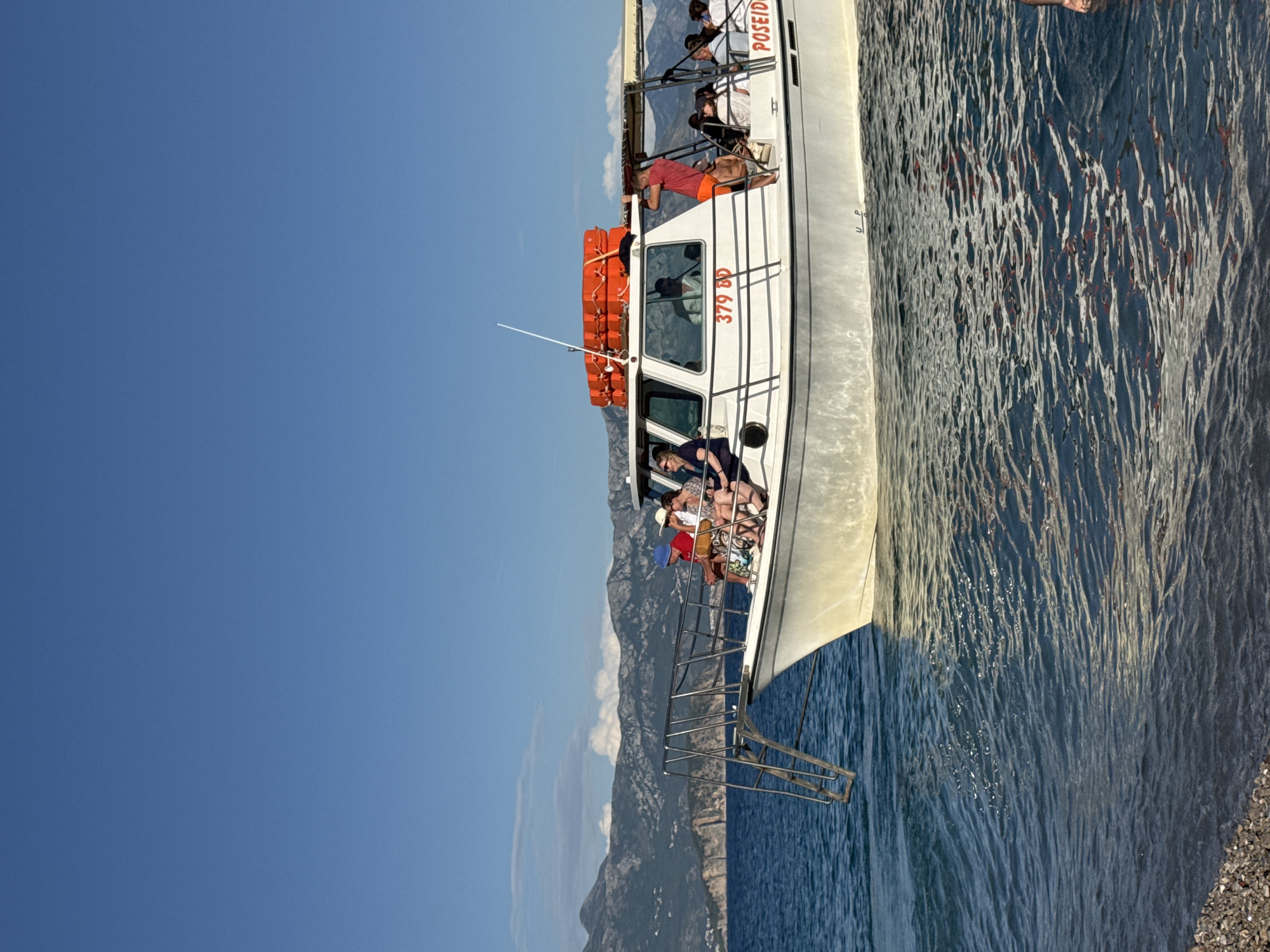 A white passenger boat named "POSEIDON" is anchored in the clear blue waters off the coast of Prijevor, Montenegro. The boat is filled with people enjoying the sunny day. In the background, lush green mountains rise majestically against a clear blue sky, suggesting a picturesque coastal landscape. The water is calm, with small waves gently lapping against the shore. The boat appears to be a tour boat, possibly offering scenic excursions along the Montenegrin coast. The scene depicts a typical tourist activity in a beautiful natural setting.