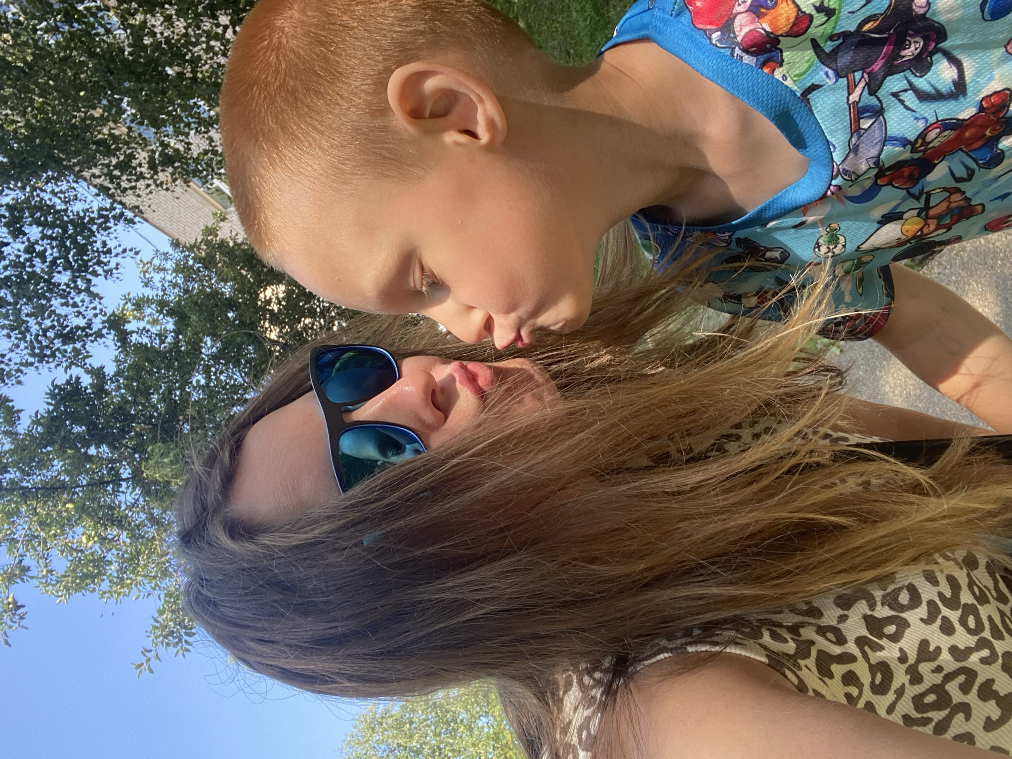 The image shows a close-up, overhead shot of two people, likely a mother and son, sharing a tender moment outdoors. The setting appears to be a sunny day in Noviy, Russia, with lush green trees and a bright blue sky visible in the background, suggesting it is likely daytime.
The foreground is dominated by the faces of the two individuals. A young boy with short, light brown hair is leaning in towards the camera, puckering his lips as if to give a kiss. He is wearing a blue t-shirt adorned with a colorful cartoon print. Partially visible below him is the face of an adult, likely a woman, with long, light brown hair that cascades around her. She is wearing dark sunglasses with blue lenses, and her lips are also slightly parted, suggesting she is either kissing the boy back or about to. Her upper body is clad in a patterned top with a leopard-print design.
The overall emotion conveyed is one of affection and warmth. The close proximity of the subjects and their facial expressions indicate a loving interaction. The bright sunlight casts gentle shadows, highlighting the contours of their faces and hair, and contributing to the cheerful, outdoor atmosphere. No text is visible in the image.