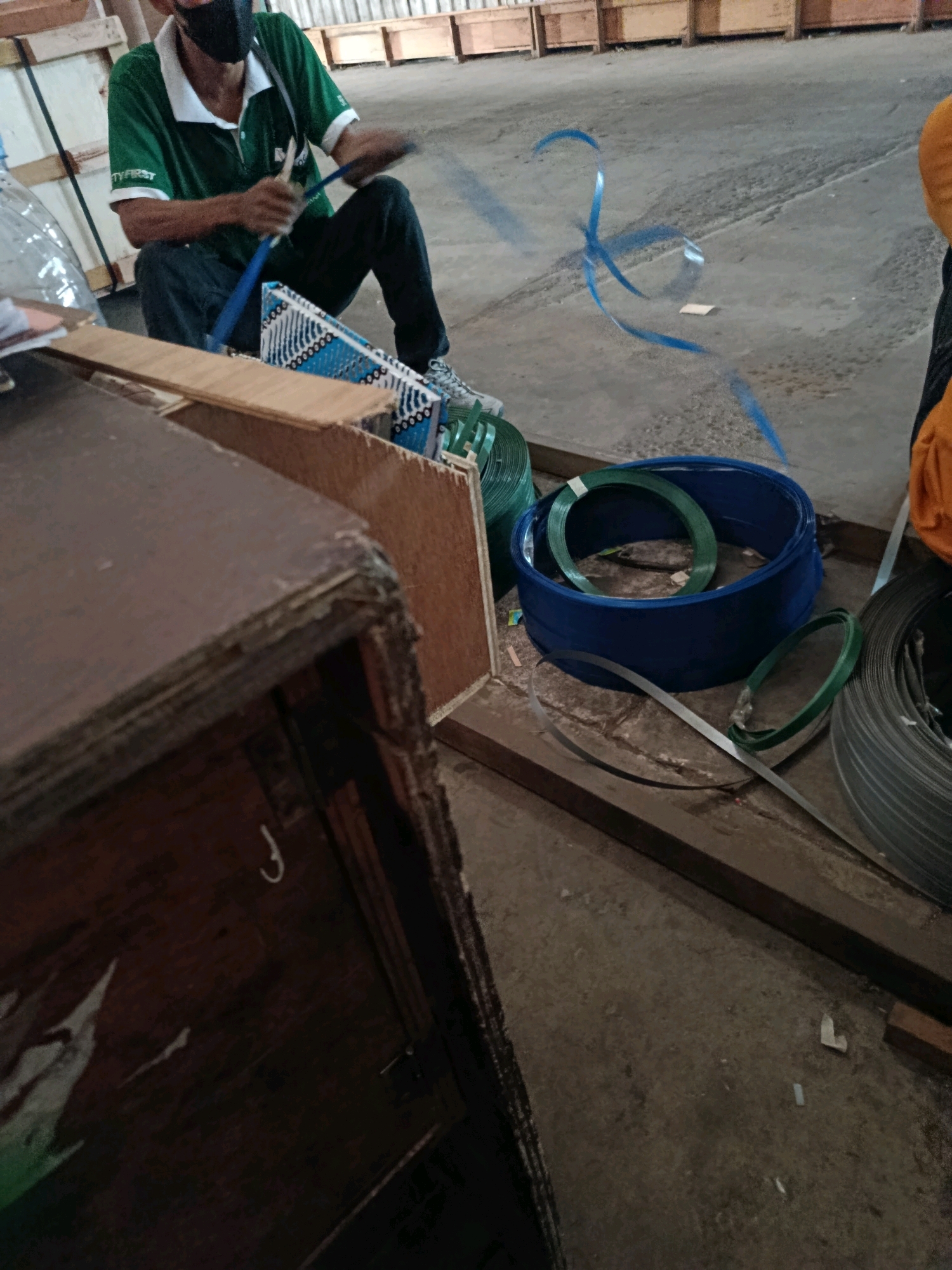 In Kembangarum, Indonesia, a person wearing a green shirt, dark pants, and a black face mask is kneeling on a concrete floor. The person is holding a blue strap in their hands, and it appears to be in the process of being secured around a white and blue patterned object. To the right of the person are several rolls of strapping material, including a large blue plastic container holding coiled green strapping, a smaller coil of green strapping, and a coil of dark grey strapping. In the foreground, a section of a dark brown wooden crate is visible. The background shows rows of wooden crates or shelving units. The overall environment suggests an industrial or warehouse setting.
