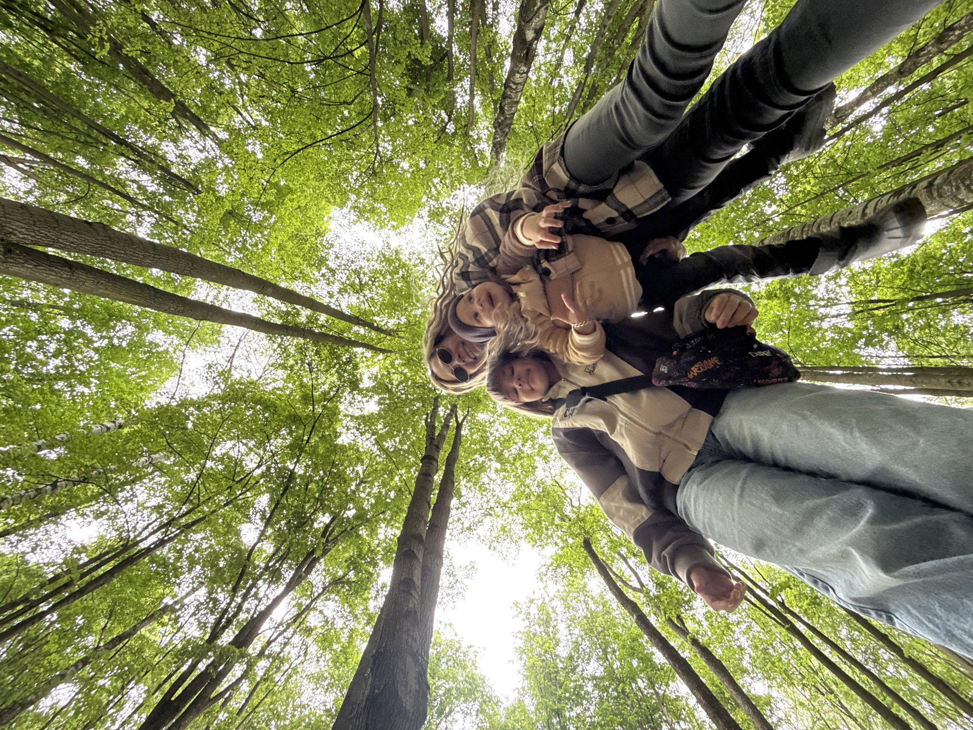 From a low-angle perspective, a group of four people is pictured beneath a canopy of tall trees in a forest. The location is Rusanovo, Russia. Three individuals are visible from the waist up, posed closely together as though they are standing together. A fourth person is visible from the waist down. The backdrop consists of trees with green leaves, creating a dense forest environment.
