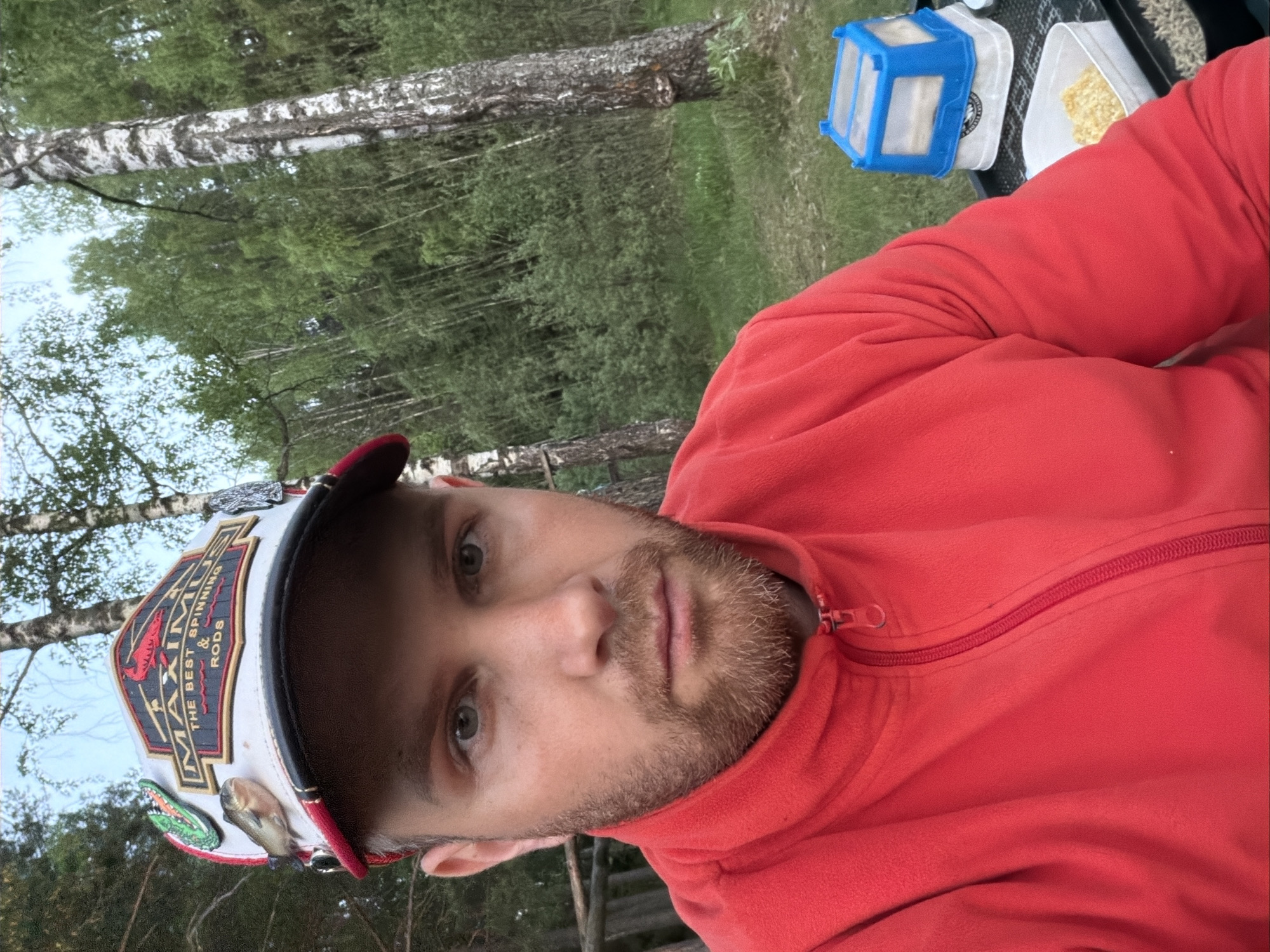 A person is pictured outdoors. The individual has light skin, a beard, and is wearing a red fleece jacket and a baseball cap with emblems on it. Two containers, one blue and one white with yellow contents, are visible. The background includes birch trees and green vegetation. It is not possible to determine if this image was taken in Chaykovsky, Russia.