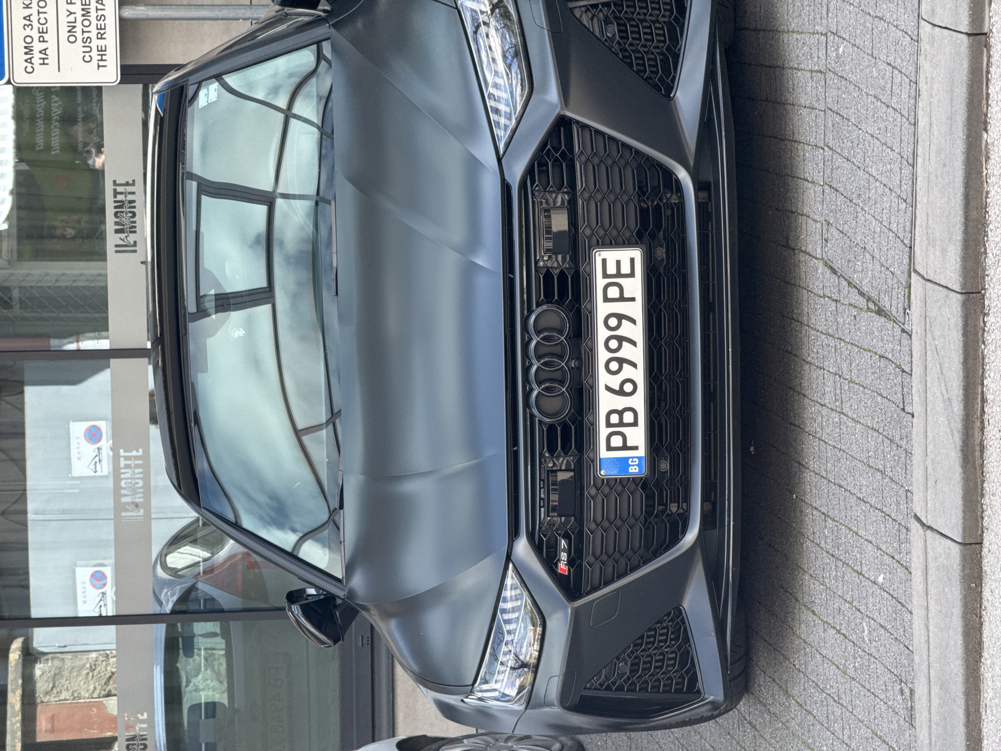 The image depicts the front of a matte black Audi RS7 with a Bulgarian license plate reading "PB 6999 PE," registered in Plovdiv ("PB"). The vehicle is parked near a building with a sign that reads "IL MONTE," possibly a restaurant, as suggested by the adjacent sign stating "САМО ЗА КЛИЕНТИ НА РЕСТОРАНТА" (ONLY FOR CUSTOMERS OF THE RESTAURANT). The location appears to be urban, with visible paving stones. The reflection in the car's windshield shows the surrounding environment.