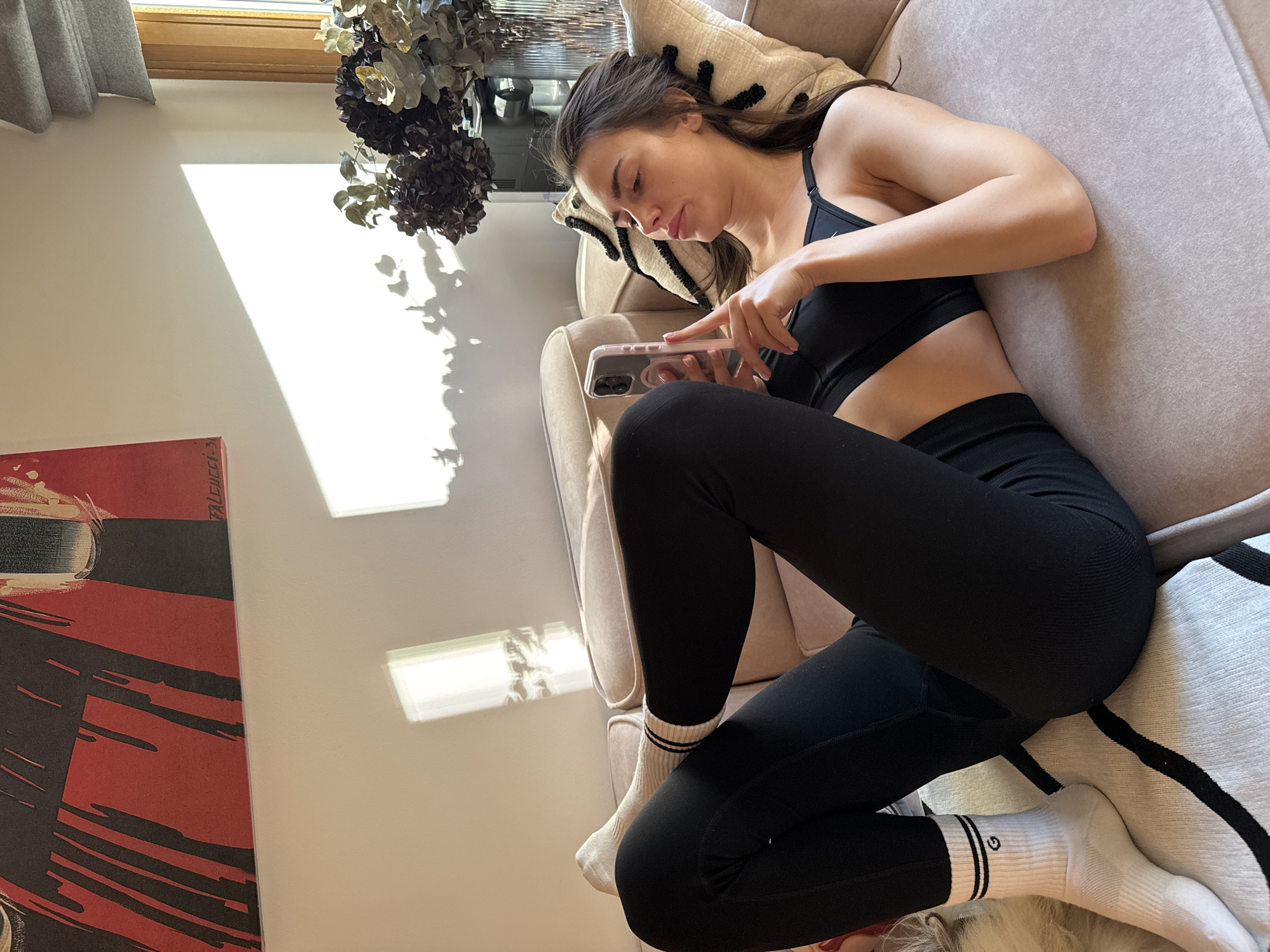 A woman is lying on a light-colored sofa in a well-lit room. She is wearing a black sports bra and black leggings, with white socks. She is holding a smartphone in her hands and looking at it. There is a window in the background, and a piece of artwork is on the wall to the left. The room appears to be in a residential setting, with decorative plants near the window.