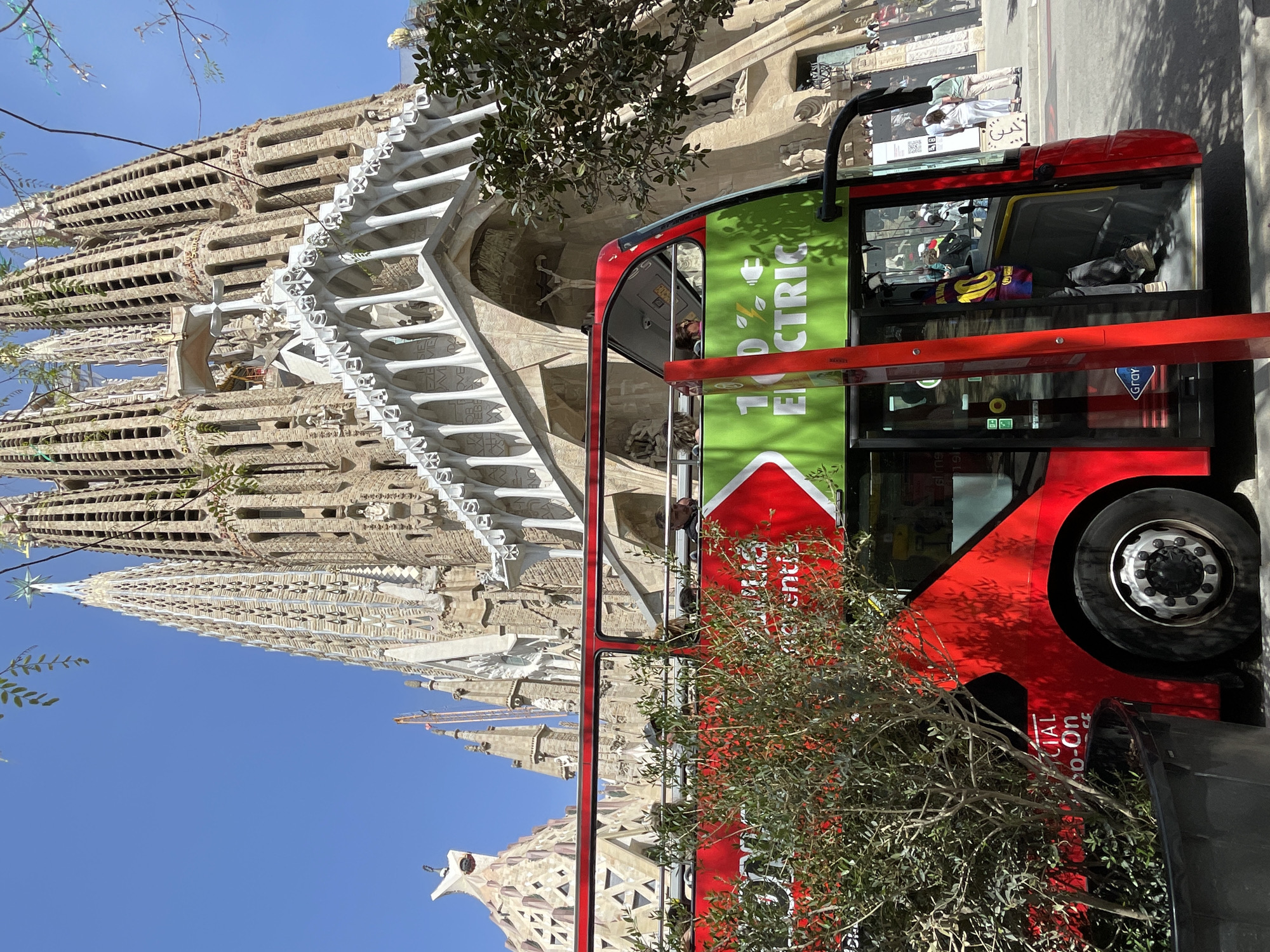 The image, rotated 90 degrees counter-clockwise, depicts a bright, sunny scene in Barcelona, Spain, featuring the iconic Sagrada Familia cathedral and a modern tour bus.
On the left, the Sagrada Familia stands prominently, showcasing its highly detailed and intricate stone architecture with multiple towering spires and ornate facades. The unique, organic forms of the building are visible, with carved figures and complex patterns adorning its surfaces. A star-shaped ornament tops one of the spires against the clear blue sky.
On the right, a red double-decker bus is positioned, partially obscured by green foliage from a tree. The bus appears to be a tourist vehicle, with passengers visible through its large windows. A large green panel on its side prominently displays the text "100% ELECTRIC," accompanied by a lightning bolt and plug icon. Other visible text on the bus includes "GRAY" and partially "Official Hop-On Hop-Off," suggesting it belongs to a tour company. Inside the bus, a colorful garment, possibly a soccer jersey with the number "10," is draped over a seat.
In the background, behind the bus, people are visible walking on a sidewalk, and a street sign with a QR code and some illegible text can be seen. The bright sunlight casts distinct shadows, indicating midday or early afternoon. The overall setting is a bustling urban environment centered around a major tourist attraction.
