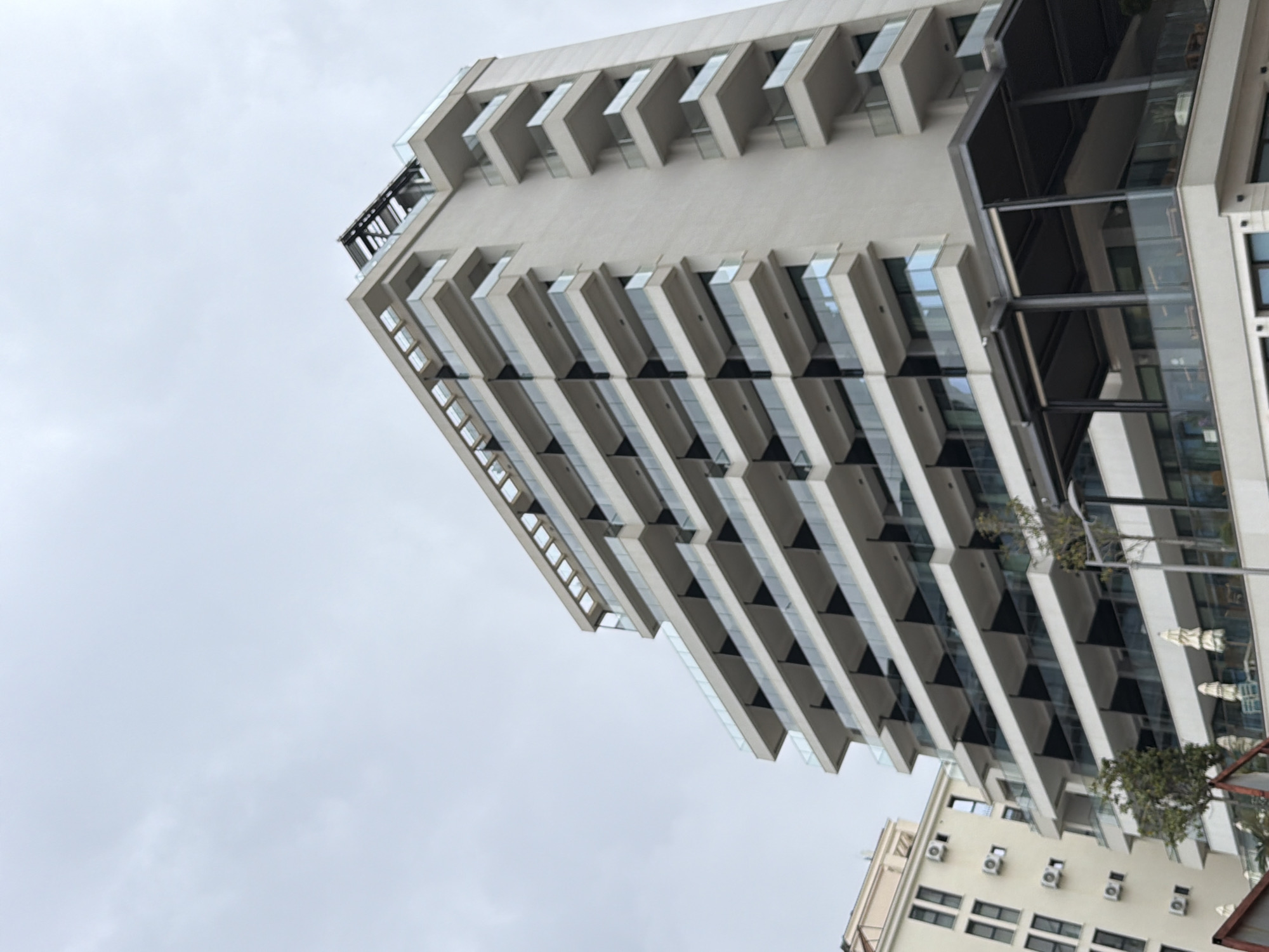 A low-angle shot of a tall, modern building, likely a hotel or residential complex, situated in Rafailovići, Montenegro. The building is characterized by its multiple floors, each featuring projecting balconies. A secondary, shorter building is visible in the lower right, and a cloudy sky dominates the background.