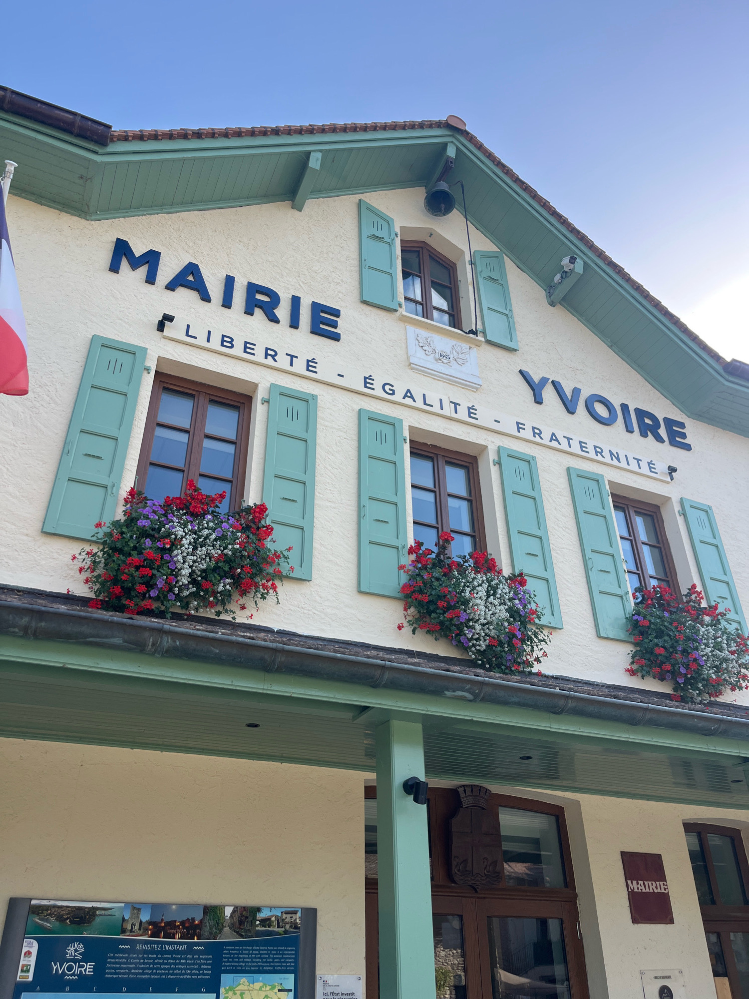 The image shows the exterior of the Mairie (Town Hall) in Yvoire, France. The building is painted a light cream color with light green shutters and trim. Above the windows, in large dark blue lettering, is the word "MAIRIE" on the left and "YVOIRE" on the right. Between these words, in smaller dark blue lettering, is the motto "LIBERTÉ - ÉGALITÉ - FRATERNITÉ". The windows are adorned with abundant flower boxes overflowing with red, purple, and white flowers. A French flag is visible on the far left. Below the main building, a sign displays historical images and text about Yvoire. A coat of arms is mounted on the wall near one of the doors. The sky is clear and blue, suggesting a sunny day. The overall scene is picturesque and well-maintained, reflecting the charm of this lakeside village.