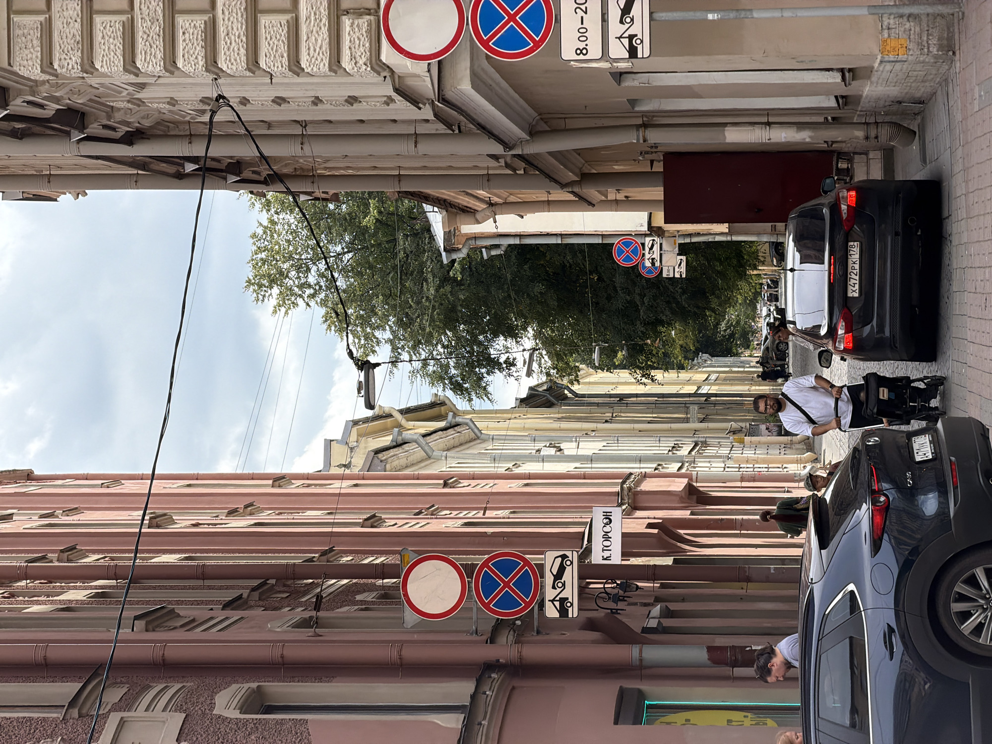 The image depicts a narrow street in Saint Petersburg, Russia, with tall, historic buildings lining both sides. The architecture features classic European styles with stucco facades, ornate details, and numerous windows. On the left, a pink building with textured walls and a light pink facade is prominent. On the right, a beige building with intricate carvings and a similar architectural style stands tall. The street itself is paved with cobblestones.
Several traffic signs are visible, including "no stopping" signs and a sign indicating that cars will be towed. One sign on the left has Cyrillic text that appears to say "К.ТОРСОН." A dark blue SUV is parked on the left side of the street, and a black sedan is parked in the center.
People are present in the scene. A man in a white t-shirt is pushing a stroller with a child in it. Another man with glasses and a beard is also visible, as is a woman in a green jacket walking away from the camera. Other pedestrians are seen further down the street. A large tree with green foliage is visible in the background, contributing to the urban landscape. The sky is cloudy. The scene appears to be during daylight hours.