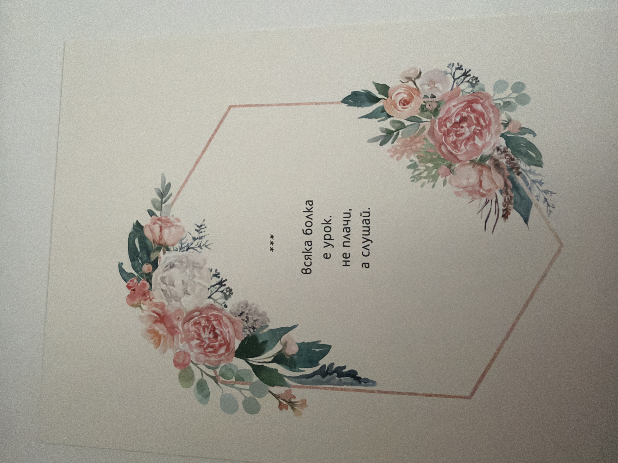 The image shows a greeting card with a floral design. The card is off-white and features a hexagonal frame made of a light pink line. On the top left and bottom right corners of the frame, there are watercolor illustrations of pink and white roses, green leaves, and small buds. In the center of the card, there is text in Bulgarian that reads:
***
Всяка болка
е урок.
не плачи,
а слушай.
This translates to:
***
Every pain
is a lesson.
don't cry,
but listen.
There is no discernible location in the city of Pazardzhik, Bulgaria depicted or implied in the image. The image is a close-up shot of the card itself.