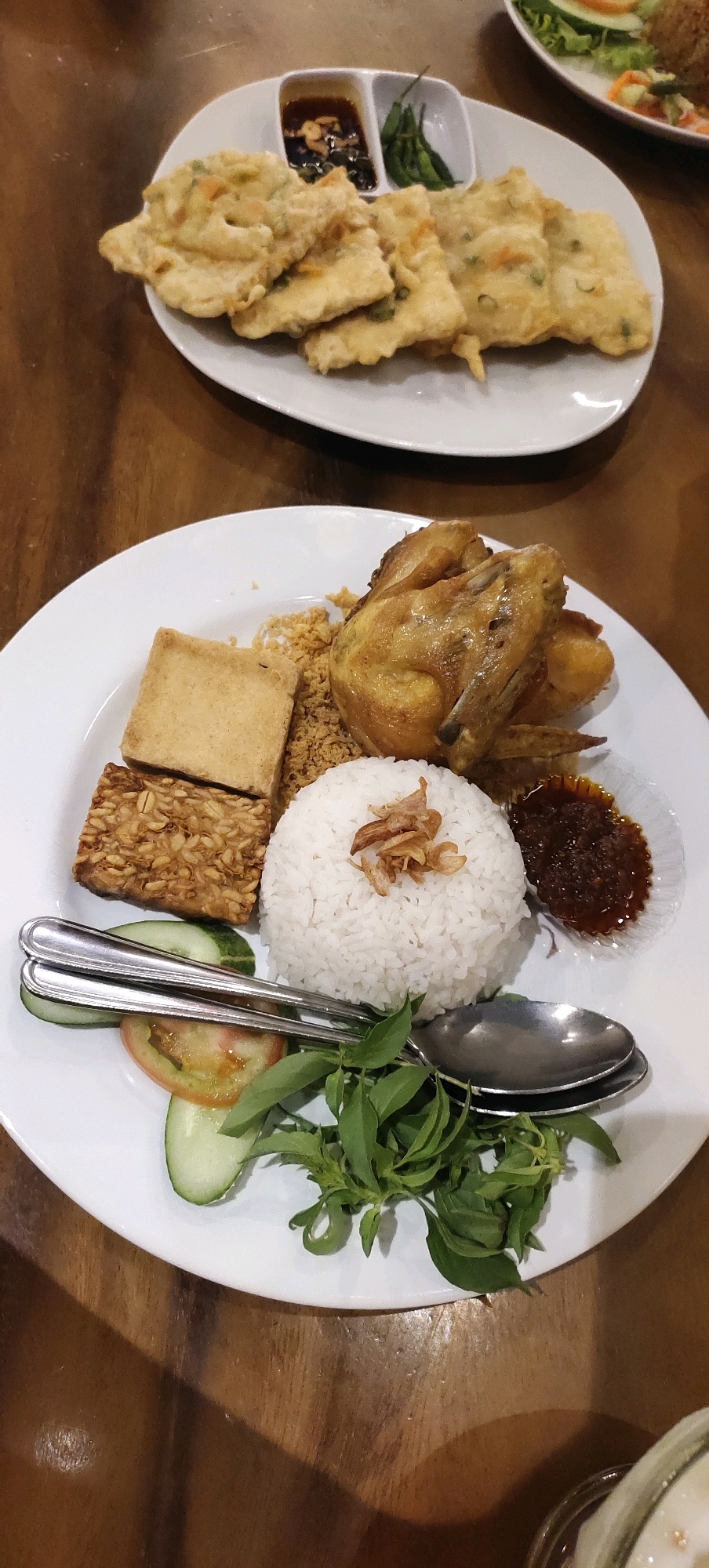 The image displays a close-up, overhead view of a white plate containing a meal. The plate is filled with white rice topped with fried shallots, a piece of fried chicken, a square of fried tofu, and a serving of fried tempeh. Accompaniments include sliced cucumber, a slice of tomato, fresh basil leaves, and a small dish of sambal. To the left of the plate are two spoons. In the background, another white plate holds fried fritters and two small dipping dishes, one with a dark sauce and the other with green chilies. The meal is set on a wooden table. The scene appears to be indoors, likely in a restaurant setting in Nganguk, Indonesia. No people are visible in the image.
