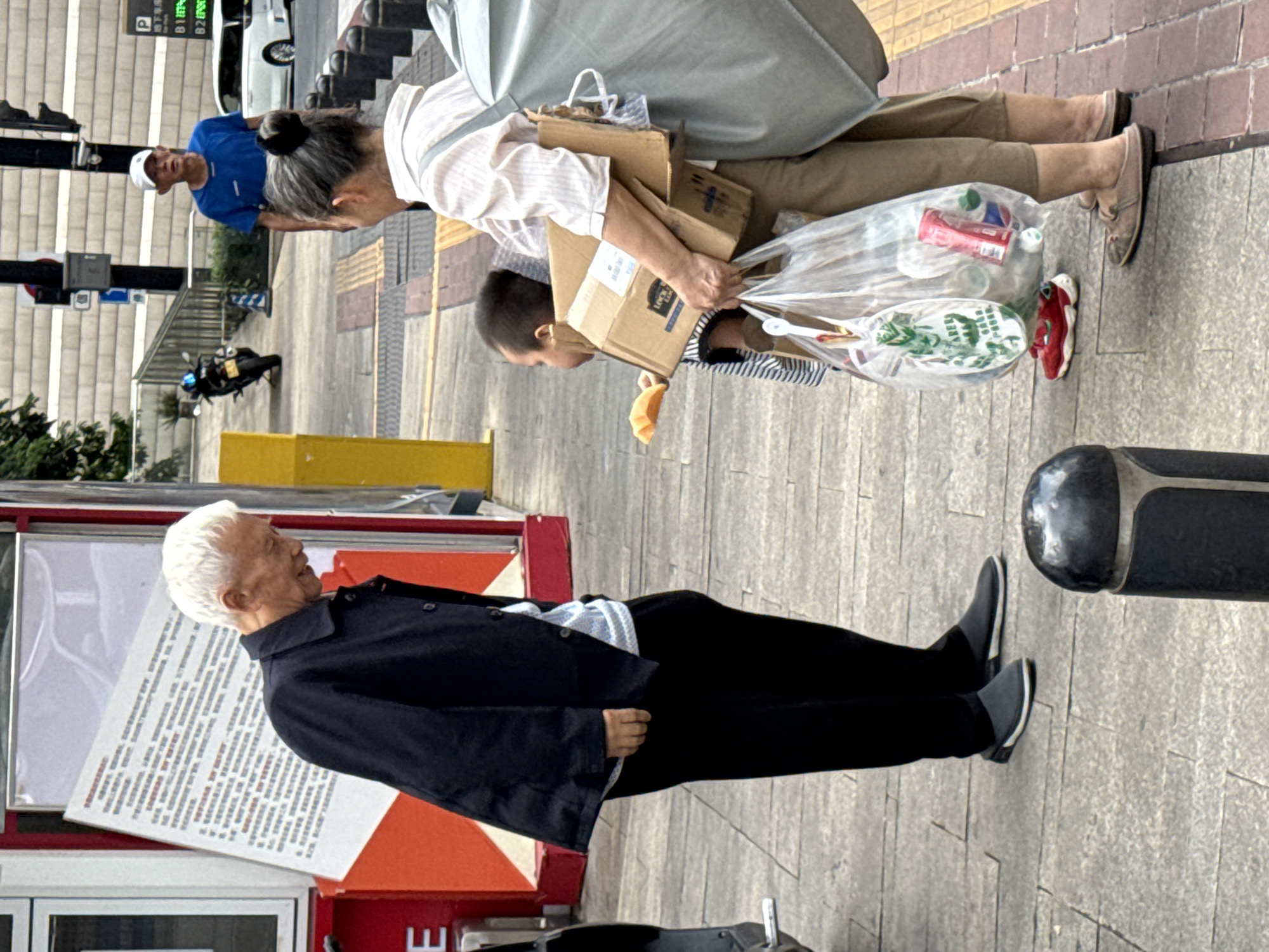 An elderly man with white hair, wearing a dark jacket over a light-colored striped shirt and black pants, stands on a sidewalk in Yuzhong District, China, facing a woman and a child. The man is leaning forward, appearing to interact with the child. The woman, wearing a light-colored striped shirt and khaki pants, carries a large gray bag over her shoulder and holds a stack of cardboard boxes in her arms. A young child, wearing a striped shirt and red shoes, stands beside the woman, holding a piece of orange food in their right hand. The child is reaching towards the elderly man. In front of the woman and child is a clear plastic bag filled with various items, including cans and bottles. The sidewalk is paved with gray stones, and a yellow bollard is visible in the background. A red and white structure, possibly a kiosk or display, is to the left of the man. Further in the background, a motorcycle and a white car are present, along with a man in a blue shirt and white cap. The overall scene suggests a street interaction on a cloudy day.
