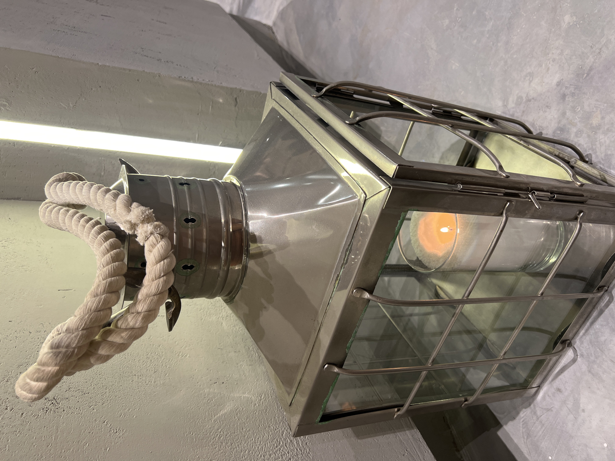An outdoor, close-up, eye-level shot shows a metal lantern with a rope handle, illuminated by a lit white candle inside. The lantern has a metallic gray finish and a glass enclosure with metal bars. It sits on a gray concrete floor. To the left of the lantern is a textured, light gray wall, and to the right, a vertical light fixture casts a bright glow on a similar gray wall. The scene is consistent with an outdoor seating area in Sochi, Russia, likely a cafe or restaurant.