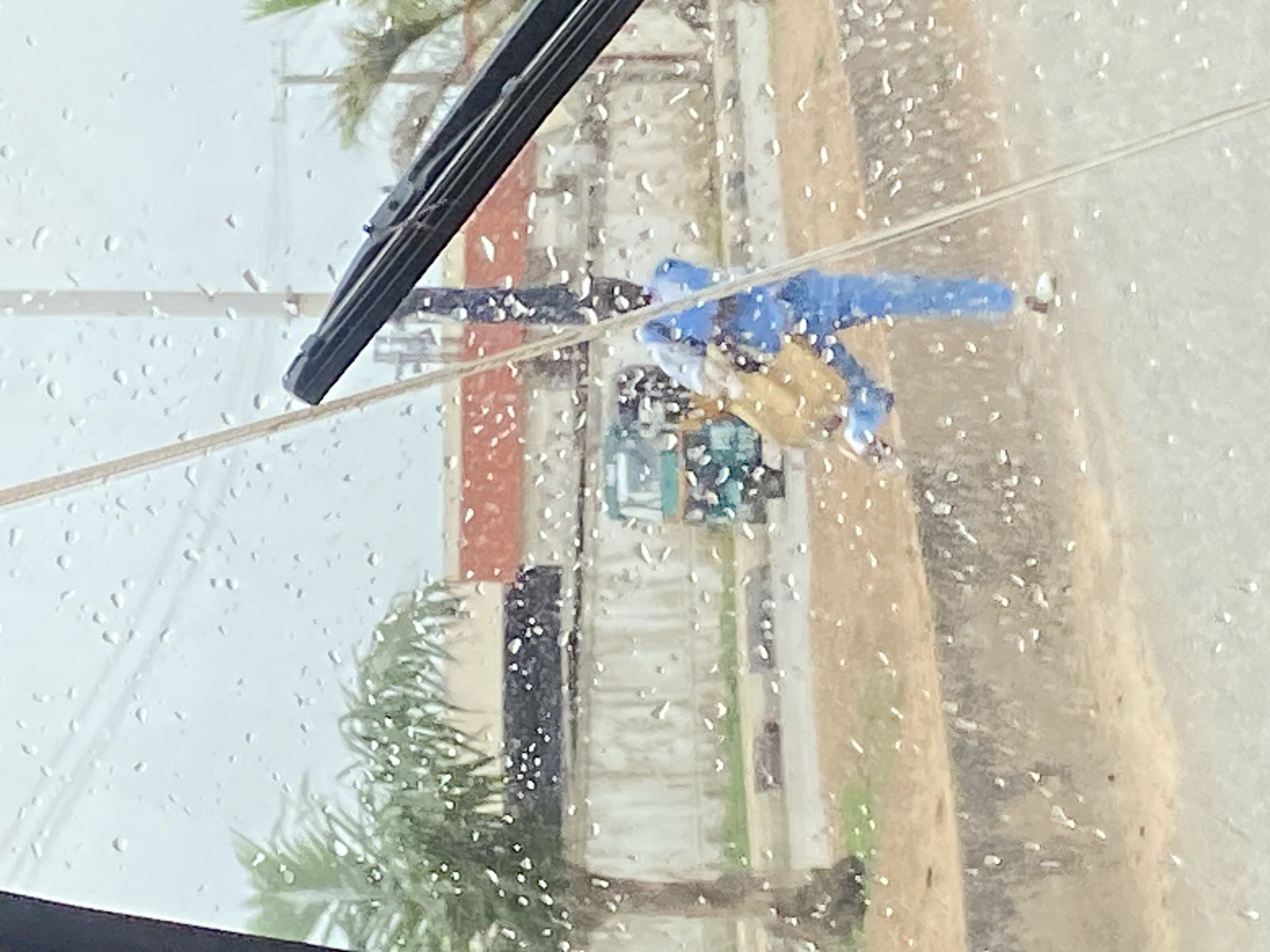 The image shows a person in a blue uniform carrying a light brown bag, walking on a street in Barakin Lerawi, Nigeria. The street is wet with raindrops, and the view is partially obstructed by raindrops on a windshield. In the background, there is a white wall with red and black sections, a palm tree, and a green and blue vehicle. The person appears to be walking away from the viewer.