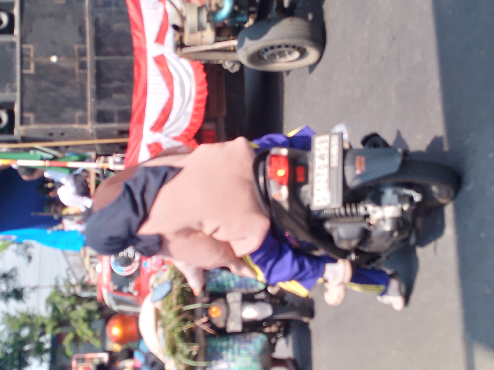 In the city of Kertosono, Indonesia, this image captures a street scene with multiple individuals and vehicles. A person wearing a pink hijab and a light pink sweater is seated on a black motorcycle, with purple pants and white shoes. They are carrying a basket of green vegetation on the back of the motorcycle. Behind them, another motorcycle is visible. In the background, a red vehicle, possibly a modified car or a three-wheeled vehicle, is present. To the right, a large set of speakers is stacked, with a red and white banner draped in front of them. The scene appears to be outdoors on a paved road, with trees visible in the upper left corner. The lighting suggests it is daytime, with shadows cast on the ground.