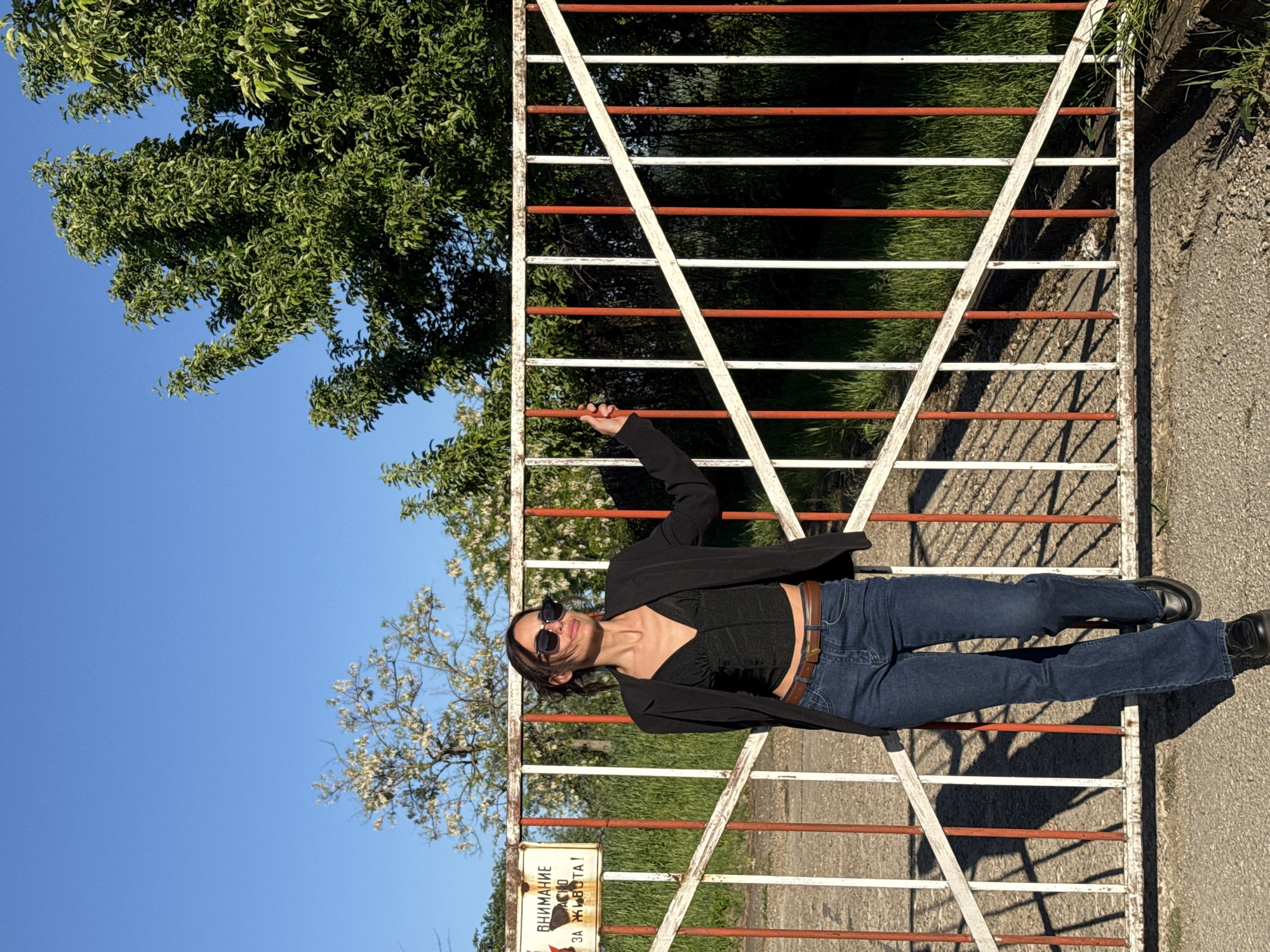 A woman poses on a metal gate in an outdoor setting, seemingly in Pleven, Bulgaria, based on the Cyrillic script visible on a sign. The gate is painted with alternating white and red horizontal bars. She wears a black blazer over a dark top, blue jeans, and black shoes. A brown belt cinches her waist. She is accessorized with sunglasses. The background features lush green trees under a clear blue sky. A paved surface, likely a road or walkway, is visible to the right.