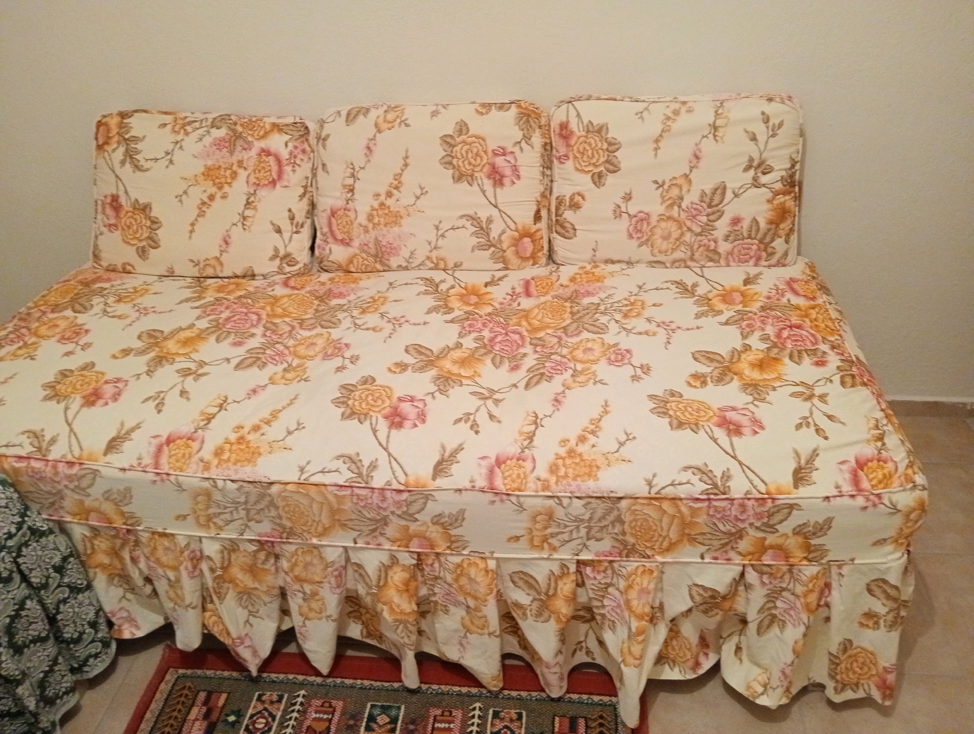 The image shows a floral-patterned sofa with three rectangular cushions. The sofa is covered in a cream-colored fabric with a repeating pattern of pink and orange flowers and green leaves. The cushions are also covered in the same fabric. The sofa has a ruffled skirt along the bottom. A portion of a dark green patterned blanket is visible on the left side of the sofa. Beneath the sofa, a rug with geometric patterns in red, green, and brown is partially visible. The background is a plain, light-colored wall. The scene appears to be an indoor setting, possibly a living room or bedroom. No people are present. The image has a neutral color palette dominated by the floral pattern of the sofa. The lighting is even and there are no harsh shadows. The image quality is moderate, with some visible pixelation. The perspective is straight on, showing the front of the sofa. Based on the available information, it is not possible to determine the specific location within the city of Silifke, Turkey.