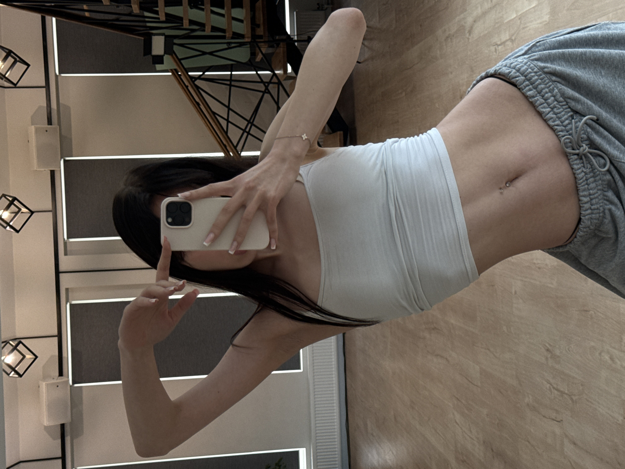 A young woman is taking a mirror selfie in a modern interior. She has long, dark hair and is wearing a white tube top and grey sweatpants. A delicate bracelet with a four-leaf clover charm adorns her wrist. Her midriff is visible, showing a navel piercing. The background includes a staircase, contemporary lighting fixtures, and wooden flooring. The lighting suggests it is daytime. The overall mood is casual and relaxed. There is no visible text in the image.
