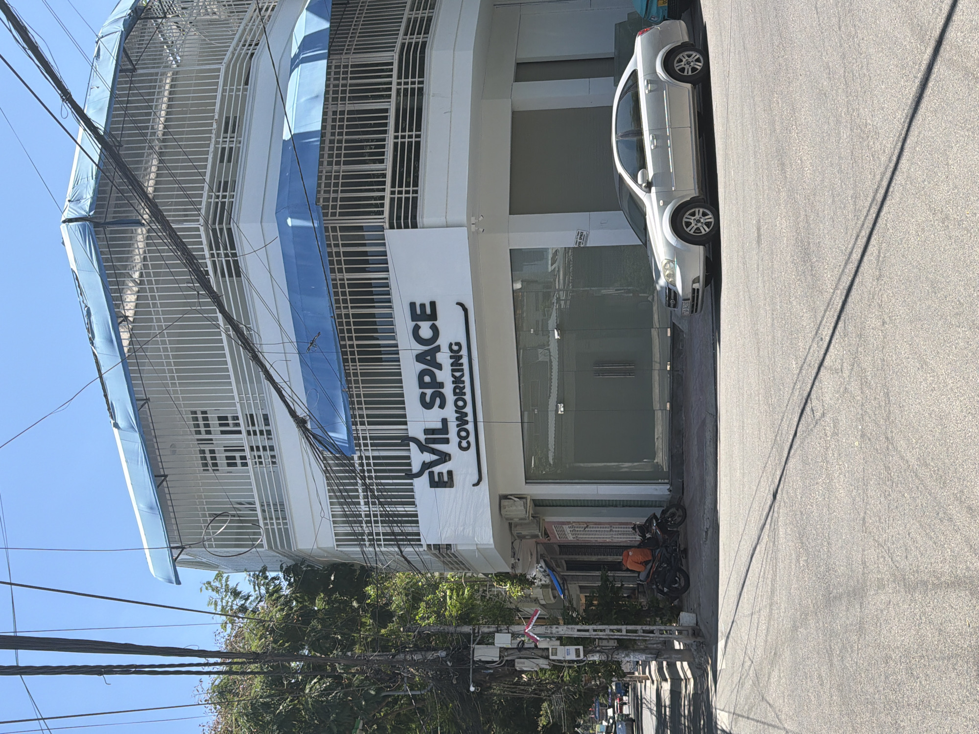 This is a street-level, eye-level, medium shot photograph of a multi-story building with the signage "EVIL SPACE COWORKING" on its facade. The building is white with blue awnings and is surrounded by a white metal fence. A silver sedan is parked on the street in front of the building, and a motorcycle with an orange-clad individual is parked on the sidewalk to the left. The street is paved, and trees and utility poles with numerous wires are visible along the left side of the frame. The sky is clear and blue. The location is identified as Bac Nha Trang Ward, Vietnam.