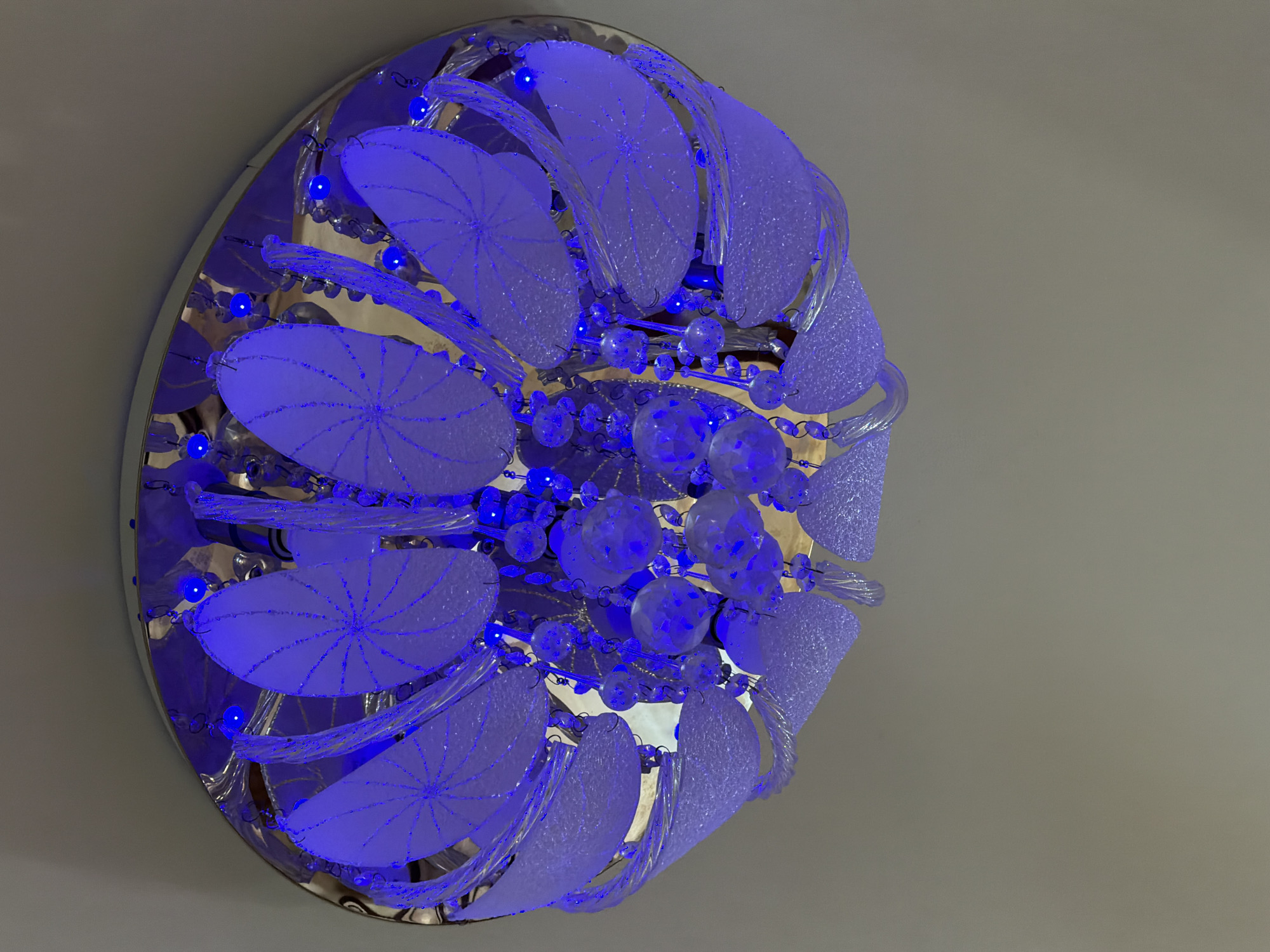 The image shows a modern, circular chandelier fixed to a neutral-colored ceiling. The chandelier features translucent, petal-shaped elements with radiating designs and strands of glass beads or crystals. Blue LED lights illuminate the structure. The location is unavailable from the visual data.
