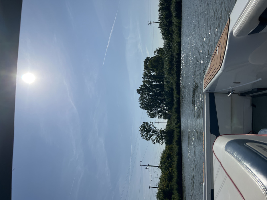 The image depicts a daytime scene from the stern of a white powerboat navigating a calm waterway. The sun is prominently visible as a bright orb in the upper-center sky, casting reflections on the water's surface. The sky is clear light blue with a few faint cirrus clouds and at least two visible airplane contrails. The opposite bank of the waterway is lined with dense green vegetation, including trees and bushes. Utility poles with overhead lines, consistent with power or railway infrastructure, are visible rising above the treeline on both the left and right sides of the background. The boat's interior, in the foreground, features white upholstered seating with dark blue accents and red piping on the left, and a brown, textured non-skid decking material on the gunwale to the right. Chrome grab handles are visible on the boat's sides. No individuals are visible. This scene is consistent with a view from the Sorraia River within the municipality of Coruche, Portugal.