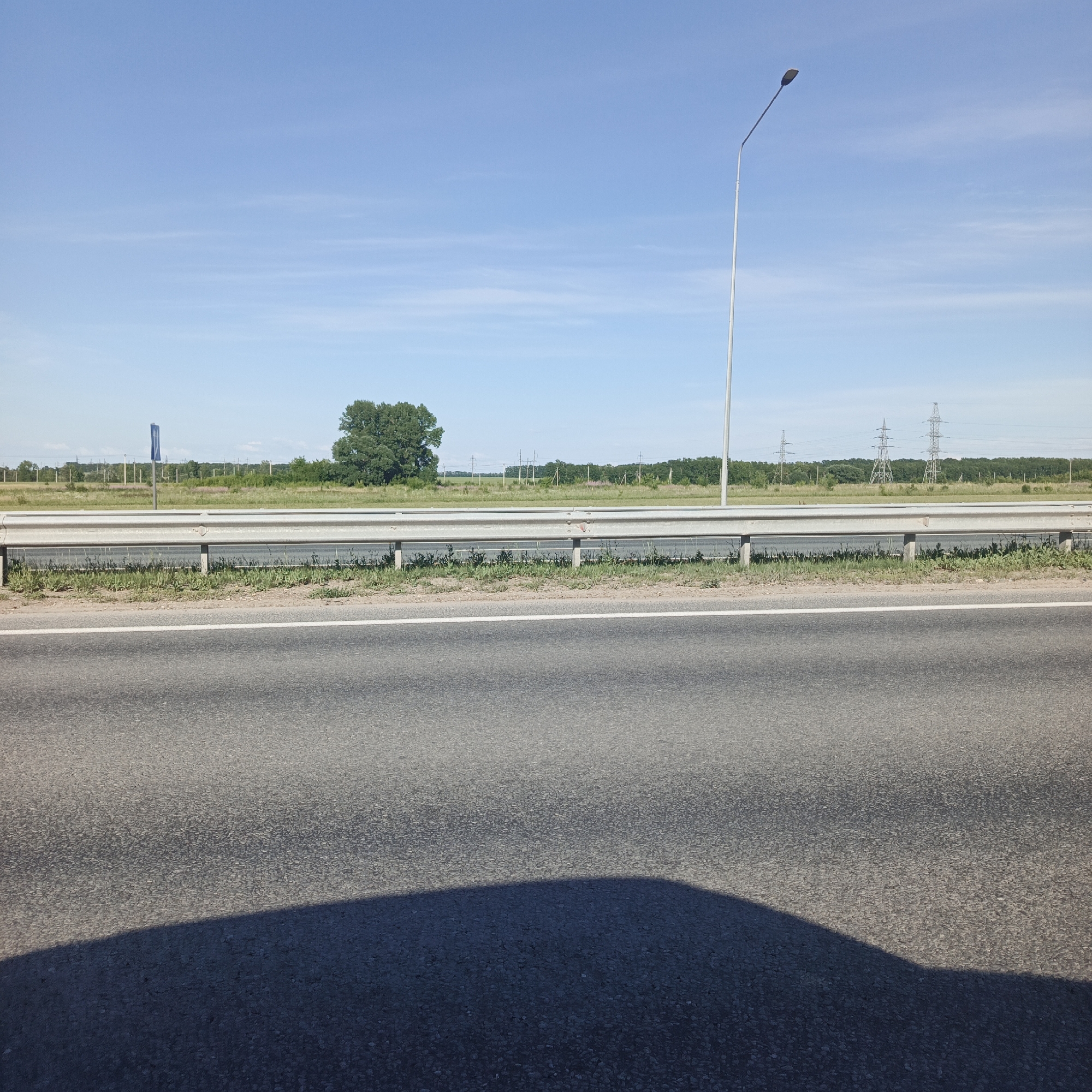 The media file shows a roadside scene in Bulgakovo, Russia, under a clear blue sky. The foreground features an asphalt road with a white line marking the lane, and a shadow cast by an unseen object, likely a vehicle, falls across the lower portion of the frame.
In the mid-ground, a metal guardrail runs along the edge of the road, separating it from a grassy embankment. Beyond the embankment, a flat landscape is visible, characterized by fields, trees, and several tall power line towers. A solitary, mature tree stands prominently on the left side of the frame, while a tall lamppost rises on the right. A blue sign is partially visible in the distance behind the guardrail.
The scene appears to be set during the daytime, with bright sunlight suggesting fair weather. The overall impression is one of a quiet, rural highway setting. There are no people visible in the frame, and the activity is limited to the static elements of the landscape and infrastructure. The presence of power line towers suggests a somewhat developed area, yet the vast sky and open fields evoke a sense of space and tranquility. No discernible text is visible in the image.