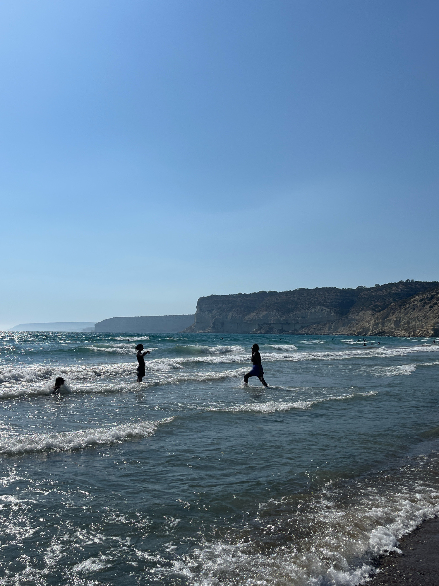 The image depicts a bright, sunny day at a beach with a clear blue sky. The sea is active with moderate waves crashing onto the shore, creating white foam. In the shallow water, several individuals are visible. In the foreground, on the left, a person is partially submerged in the water, appearing to be playing or swimming. Further to the right, another person stands in the water, facing towards the right side of the frame, with their arms raised. To the right of them, a third person is wading through the water, with one leg forward as if walking or posing. In the background, a rocky, elevated coastline stretches across the horizon, with some greenery on its slopes. The overall scene suggests a recreational activity at the seaside. The location is identified as Episkopi Beach, a coastal area within the Limassol District of Cyprus.