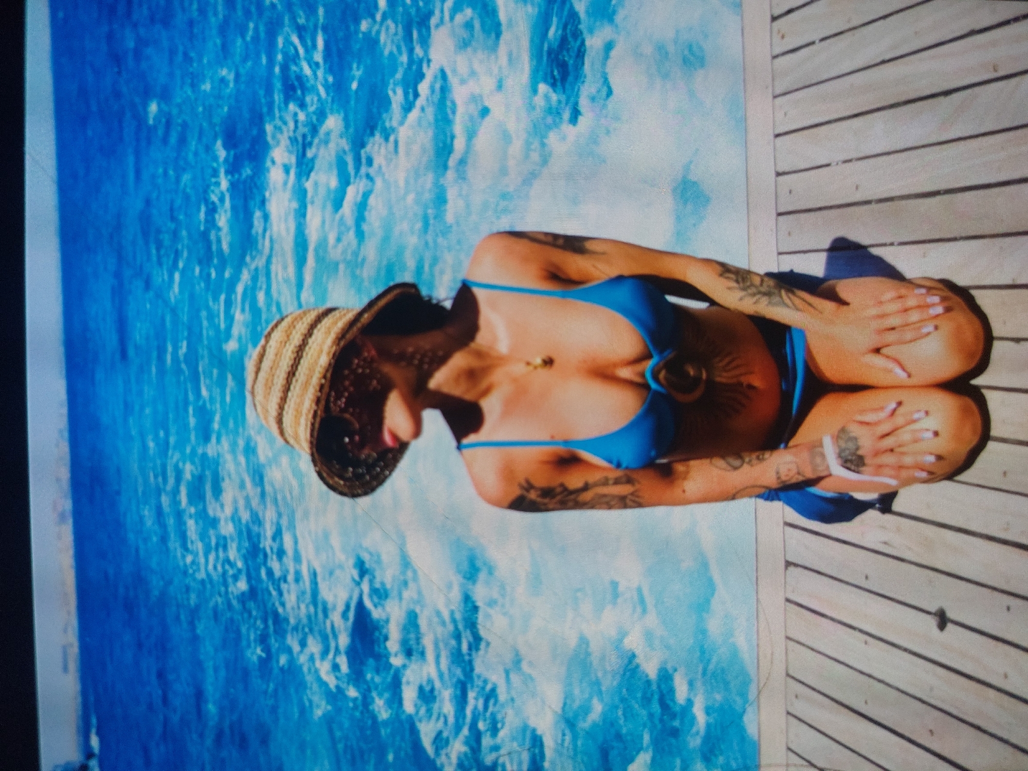 A person, likely a woman, is kneeling or sitting on their knees on a light-colored wooden deck under bright sunlight. They are wearing a vibrant blue two-piece bikini and a woven straw hat with a dark band. The individual's head is tilted downwards, partially obscuring their face with the hat. Visible details include numerous dark tattoos on both arms and perhaps the torso, as well as manicured fingernails painted a light, neutral color. A delicate gold-colored necklace with a small pendant rests on their chest.
To the left of the person is a large body of water, possibly the Black Sea, given the location context of Tsarevo, Bulgaria. The water is a vivid blue with white, foamy patterns suggesting waves or currents. The stark contrast between the bright blue water and the light wooden planks of the deck creates a striking visual. Strong shadows are cast by the person and their hat onto the wooden surface, indicating intense, direct sunlight and a clear daytime setting. The scene evokes a sense of calm and leisure by the water. No visible text is present in the image.
