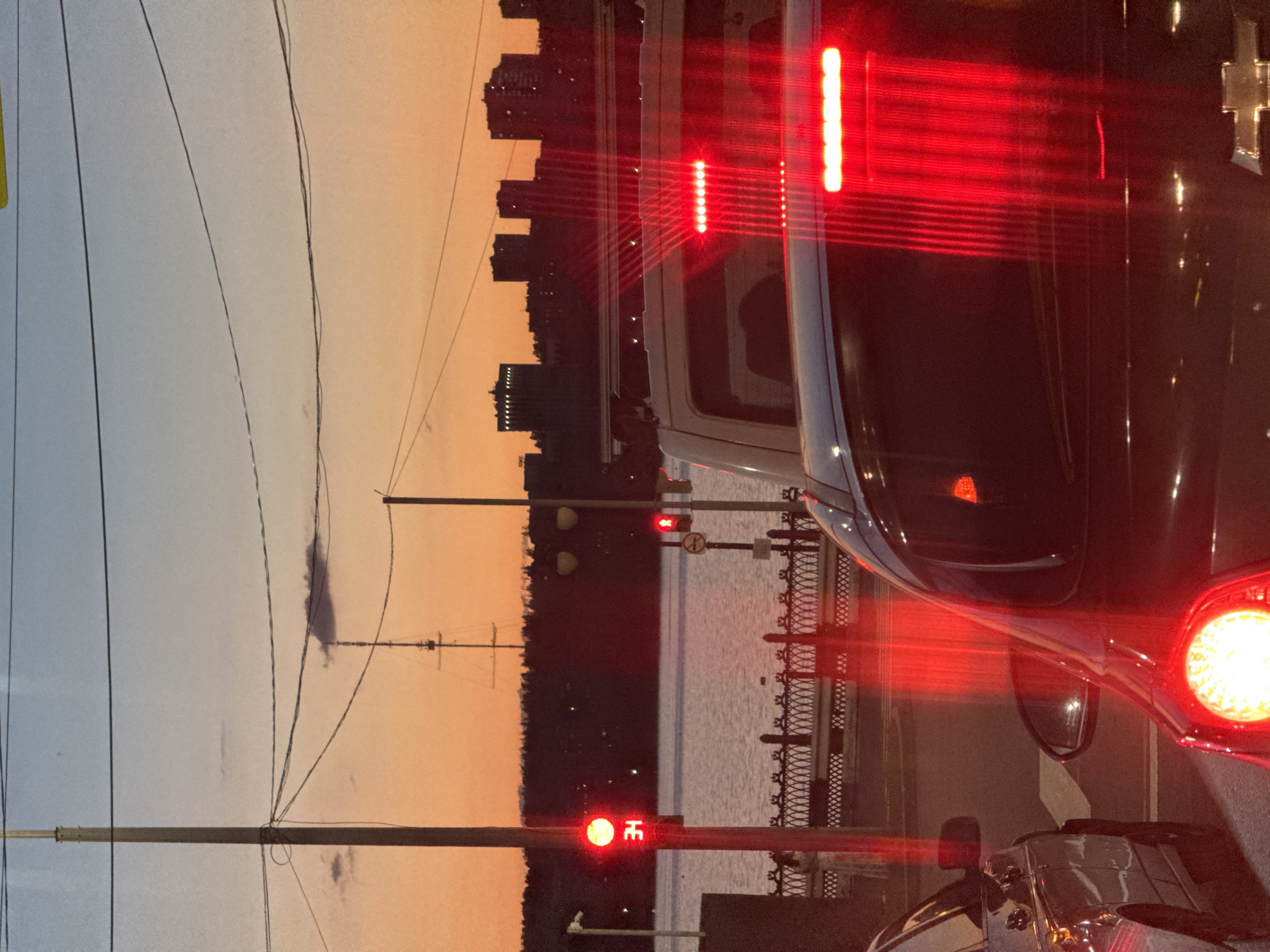 The image depicts a traffic scene at dusk in Izhevsk, Russia. A traffic light displays a red signal with the number "34" visible. In the foreground, the rear of a dark-colored car is visible, with its brake lights illuminated, casting a strong red glow. A silver car is seen to the left. In the background, across a body of water, a line of buildings and trees form the city skyline, silhouetted against an orange and blue twilight sky. A tall radio mast with rigging is prominent against the sky. Overhead power lines are also visible. The overall atmosphere suggests a moment of stillness within a typical urban environment.