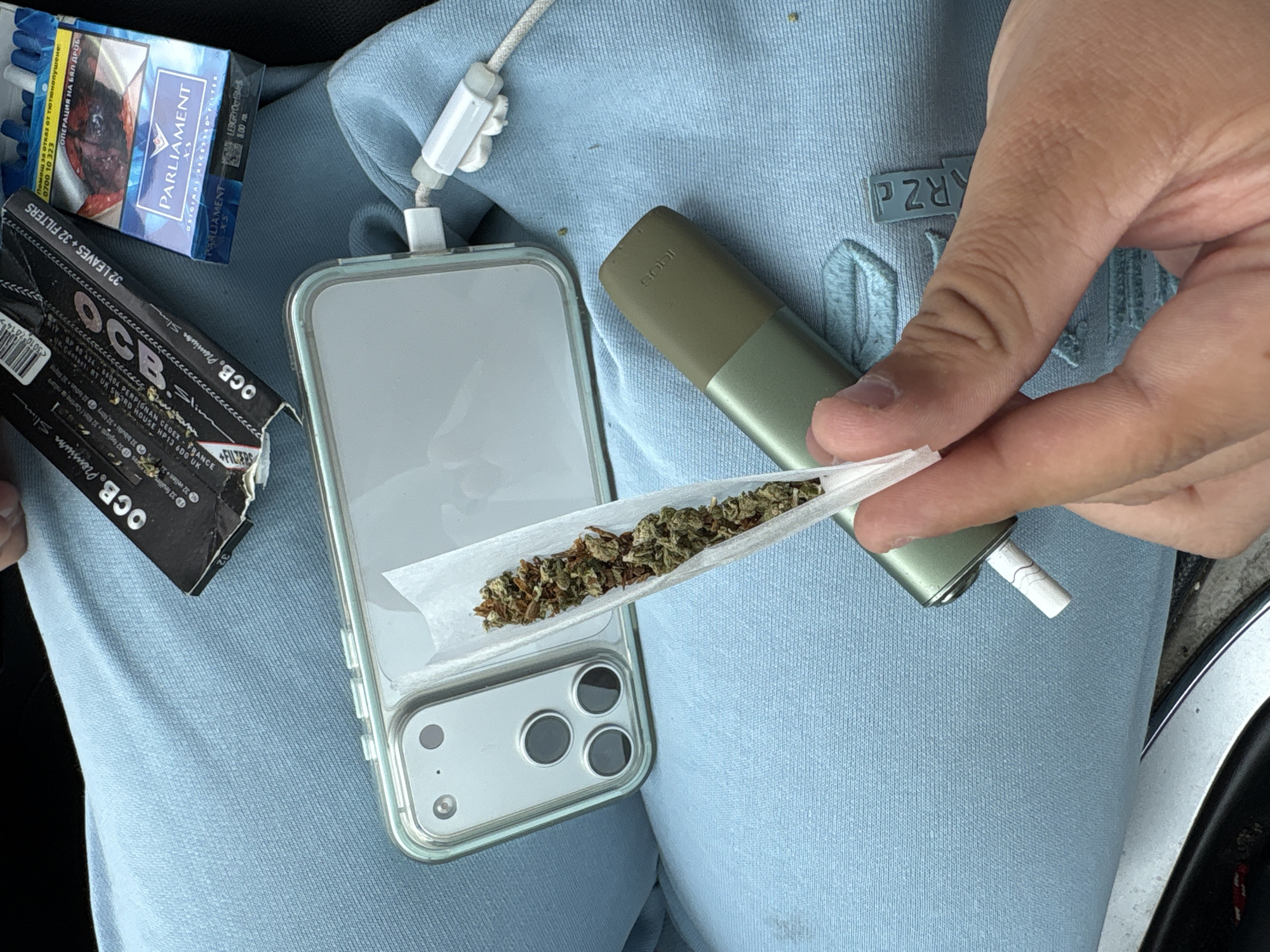 A close-up shot shows a hand rolling what appears to be cannabis in rolling paper, placed on a smartphone with a clear case. Next to the phone are a pack of OCB Premium Slim rolling papers, a pack of Parliament cigarettes, and what appears to be an IQOS device. The objects rest on a light blue fabric surface, likely clothing. The setting seems to be indoors, possibly in a car.