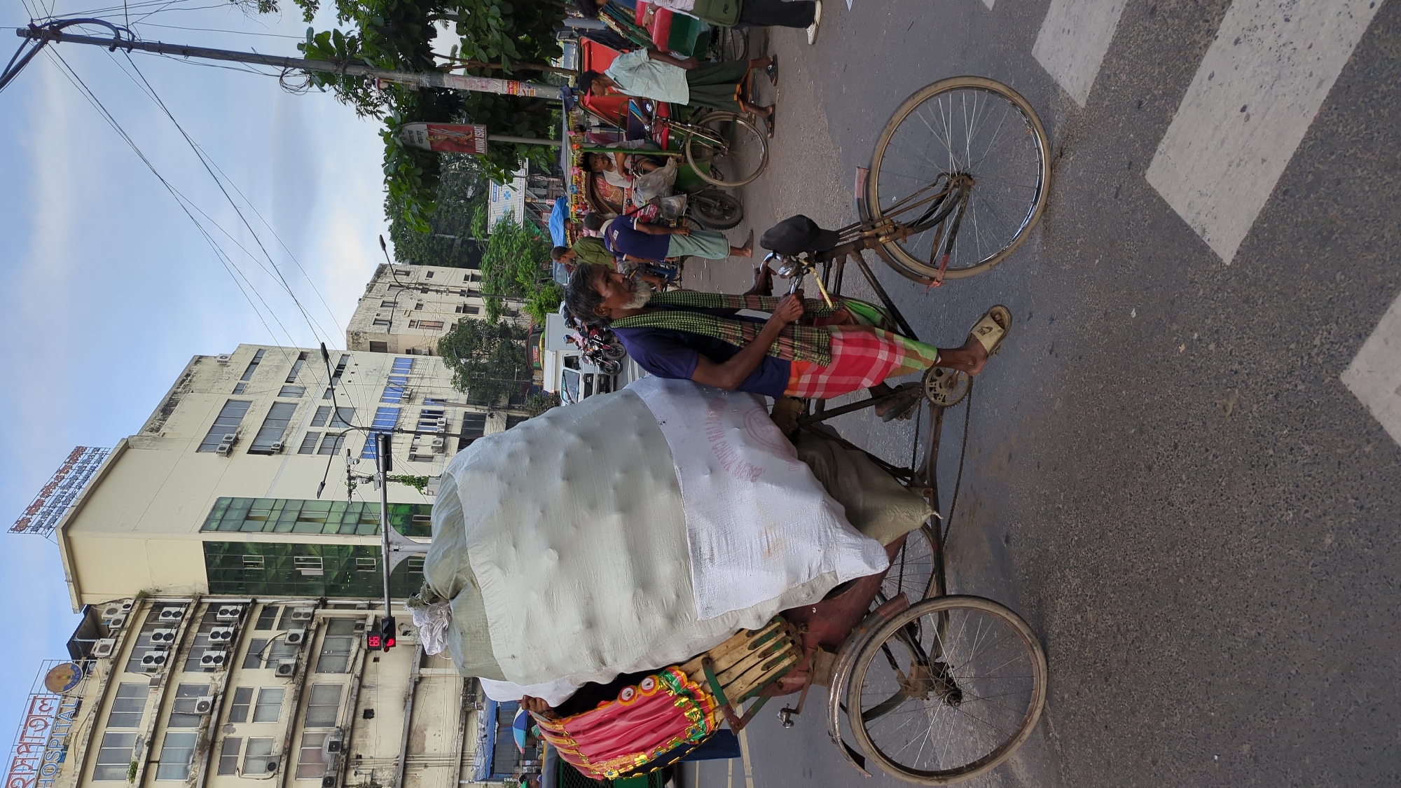 In Dhaka, Bangladesh, a rickshaw driver navigates a city street, his vehicle laden with a massive, white, woven sack. The sack, prominently displayed, is so large it dwarfs the rickshaw and the man operating it. The driver, an older man with weathered features, wears a green and brown checkered scarf around his neck and a red and white checkered lungi. He is focused on maneuvering the heavy load through traffic. In the background, a multi-story building, likely a hospital judging by the Bengali signage at the top, stands tall. Other vehicles, including motorcycles and a white van, are visible on the street, indicating the bustling nature of the urban environment. The scene is set on an asphalt road with a pedestrian crossing painted in white stripes. The sky above is overcast, suggesting a cloudy day.