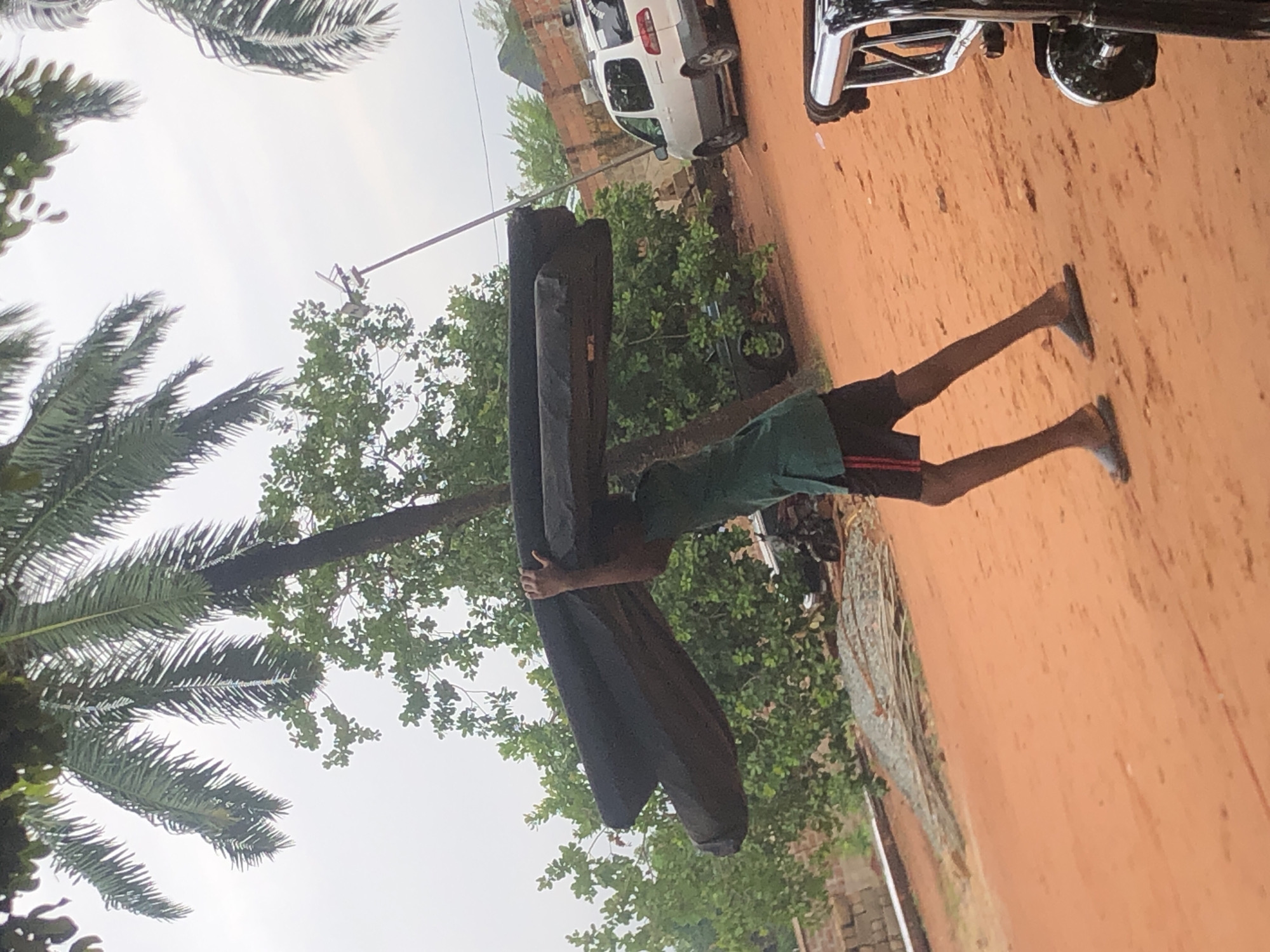 A young person in Nteje, Nigeria carries a large, dark, rectangular object, possibly mattresses, across their shoulders. They are wearing a green t-shirt and dark shorts. They are walking on a reddish-brown dirt road. To their right is lush, green vegetation and what appears to be a parked white vehicle. Palm trees are visible in the background. A motorcycle is parked nearby. The sky is overcast.