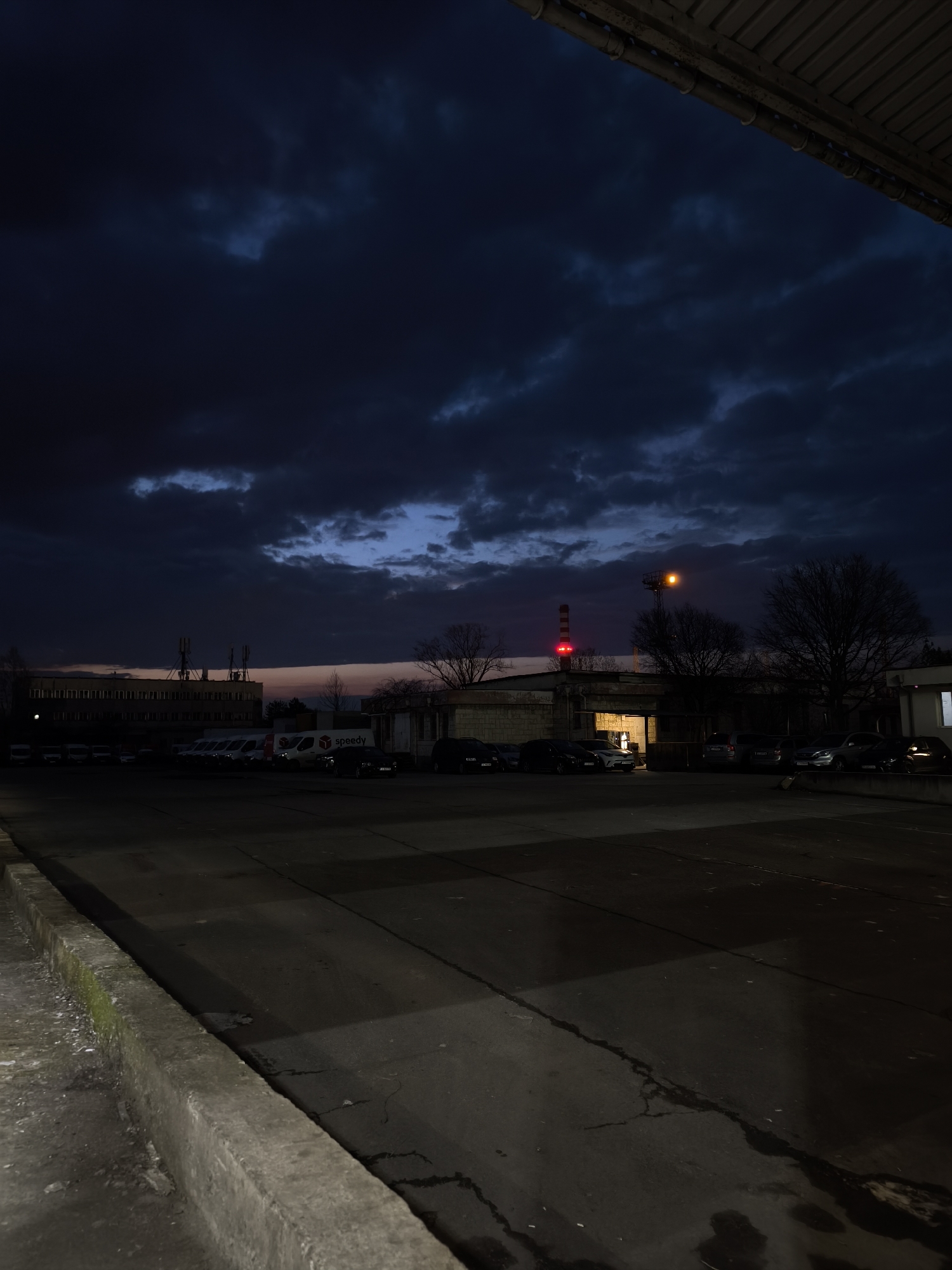 The image depicts an exterior nighttime scene in an urban setting, likely in Burgas, Bulgaria, given the presence of vehicles with "Speedy" branding, a popular courier company in Bulgaria. The foreground shows a concrete curb and a cracked asphalt parking area. In the midground are rows of parked cars and vans near a low building with a chimney and a tall industrial tower. The building appears to have an open bay or loading dock area with visible interior lighting. A red light is visible atop the chimney. The sky is overcast with dark clouds, suggesting dusk or early evening. The overall impression is of a quiet, industrial or commercial area at twilight. No people are visible in the image.