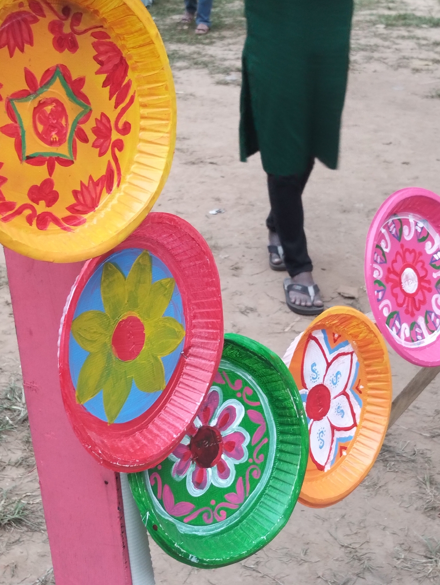 This close-up shot captures a vibrant display of handcrafted decorative items, likely at an outdoor market or fair in Dhaka, Bangladesh. The foreground is dominated by several colorful circular objects, appearing to be painted paper plates or similar lightweight discs, arranged on a pink wooden post or stand.
Each disc is uniquely adorned with intricate floral and geometric patterns in bright, contrasting colors such as red, yellow, blue, green, pink, orange, and white. For instance, the topmost disc is predominantly yellow with red stylized lotus flowers and a central green five-pointed star containing more red details. Below it, a red disc features a prominent yellow-petaled flower with a red center against a sky-blue background. Other visible discs include a green one with pink and white floral motifs, and an orange one displaying a white flower with blue accents. To the right, a pink disc showcases white and red decorative patterns. The distinctive folk art style of these decorations points to cultural significance, often associated with festivals or celebrations such typical of Bangladesh.
In the mid-ground, a person is partially visible, walking. Their lower body is seen, clad in dark pants and a dark green tunic, with open-toed sandals on their feet. The ground is unpaved, dusty, and sandy, suggesting an outdoor public area or a casual setting. The lighting is natural and soft, indicating it is daytime, possibly under an overcast sky, as there are no harsh shadows. No visible text is present in the image. The overall scene conveys a cheerful and culturally rich atmosphere.