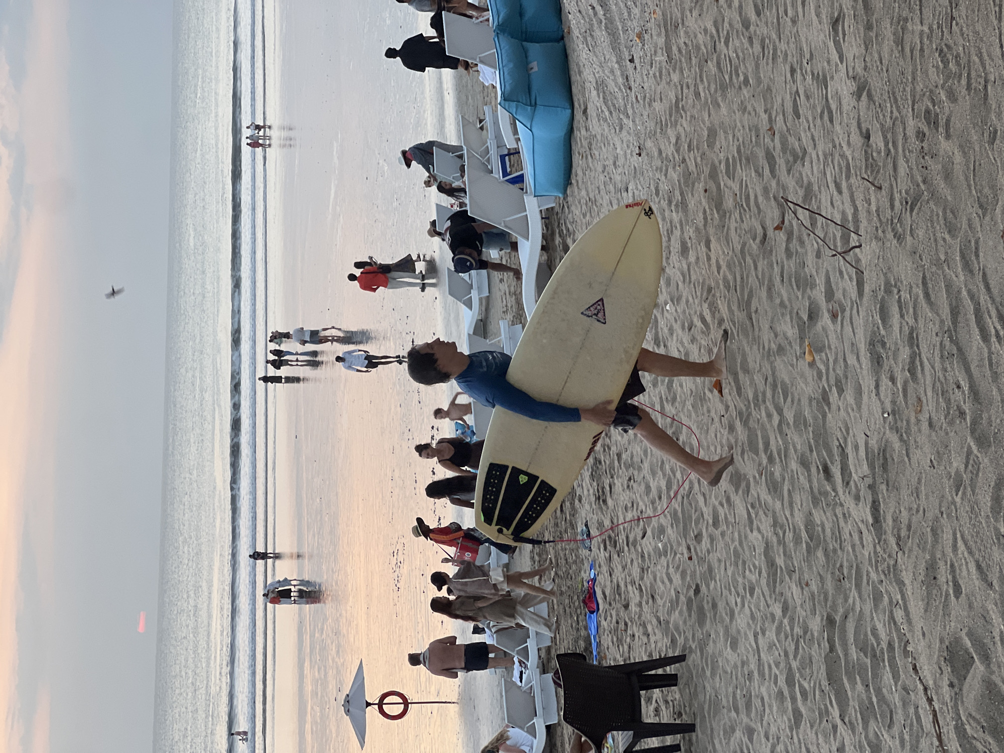 In Kuta, Indonesia, on a sandy beach during sunset, a man in a blue long-sleeved shirt carries a surfboard under his right arm. The surfboard is light-colored with black accents. He is walking across the sand from right to left in the foreground, with his legs visible from the knees down. Several beach chairs and umbrellas are scattered on the sand behind him, with people lounging and walking in the background. The ocean is visible in the distance, with gentle waves and people wading near the shore. The sky is a soft gradient of pink and orange, indicating the setting sun, with a few clouds and a bird in flight.